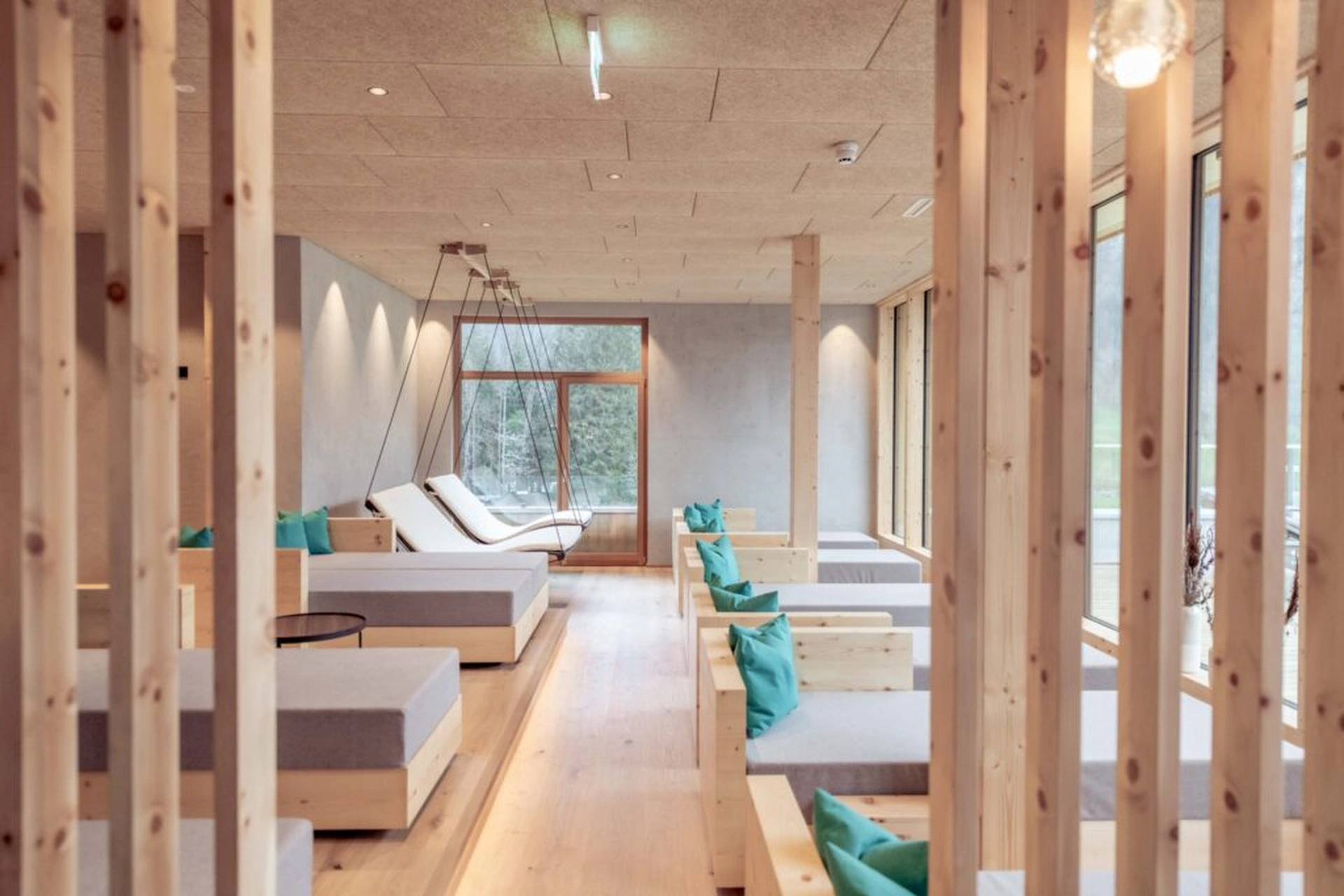 Ruheraum des Spa-Bereichs der Lechzeit in Elmen mit Zirbenholz-Liegeboxen, türkisen Kissen, hängenden Schaukeln, bodentiefen Fenstern mit Waldblick und warmem indirektem Licht.