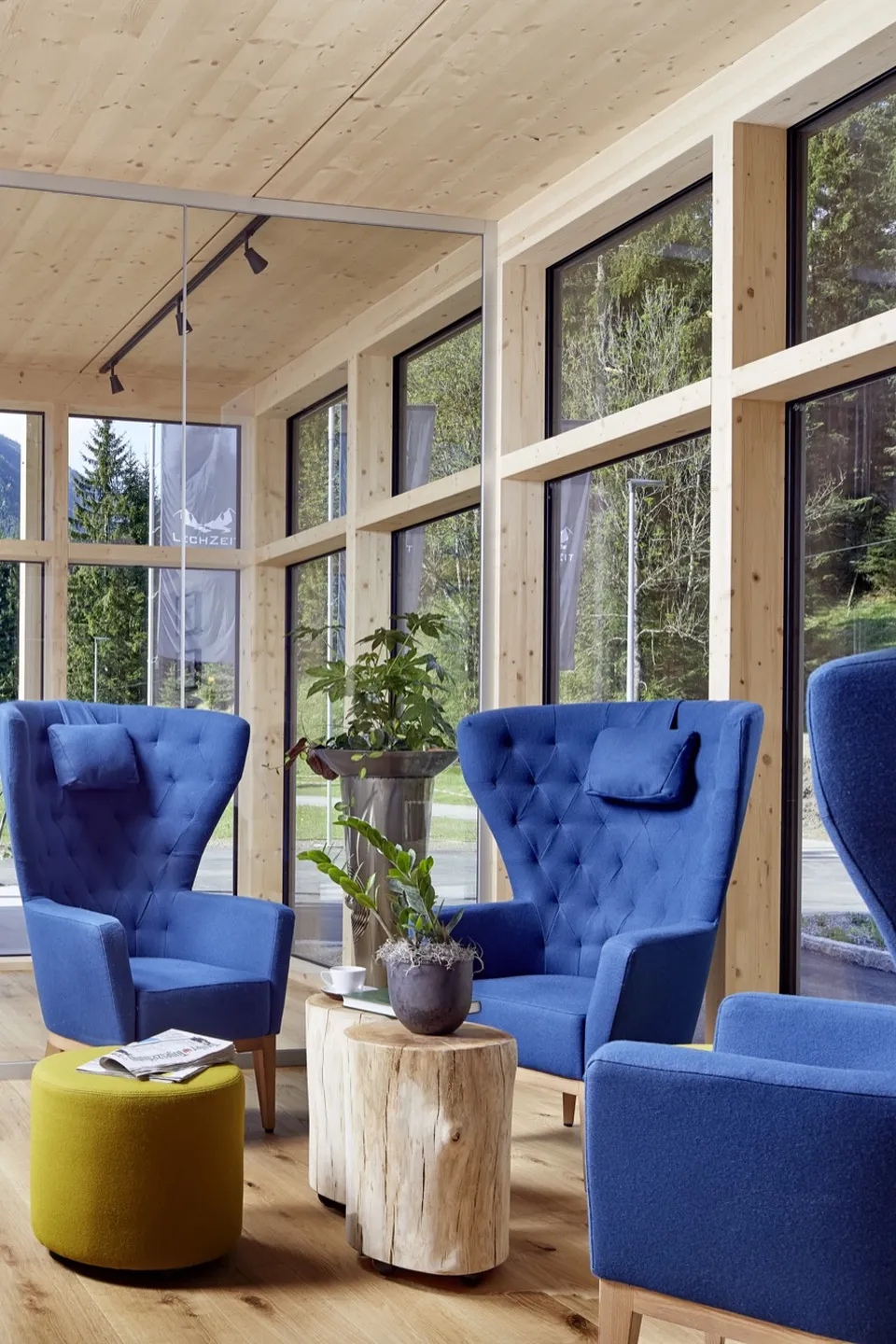 Lobbybereich des LechZeit in Elmen mit drei blauen Ohrensesseln, gelbem Pouf, Baumstumpf-Beistelltischen, Zimmerpflanze und Panoramafenstern mit Blick auf Bergwiese und Wald.