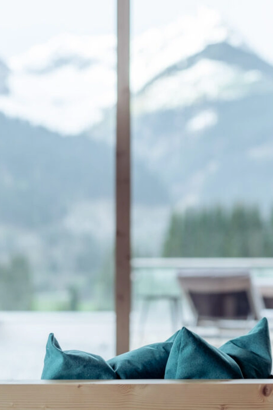 Detailaufnahme türkiser Kissen auf einer Zirbenholz-Liegebox im Spa-Bereich der Lechzeit in Elmen, im unscharfen Hintergrund Panoramafenster mit Blick auf verschneite Berggipfel und Liegestühle auf der Terrasse.