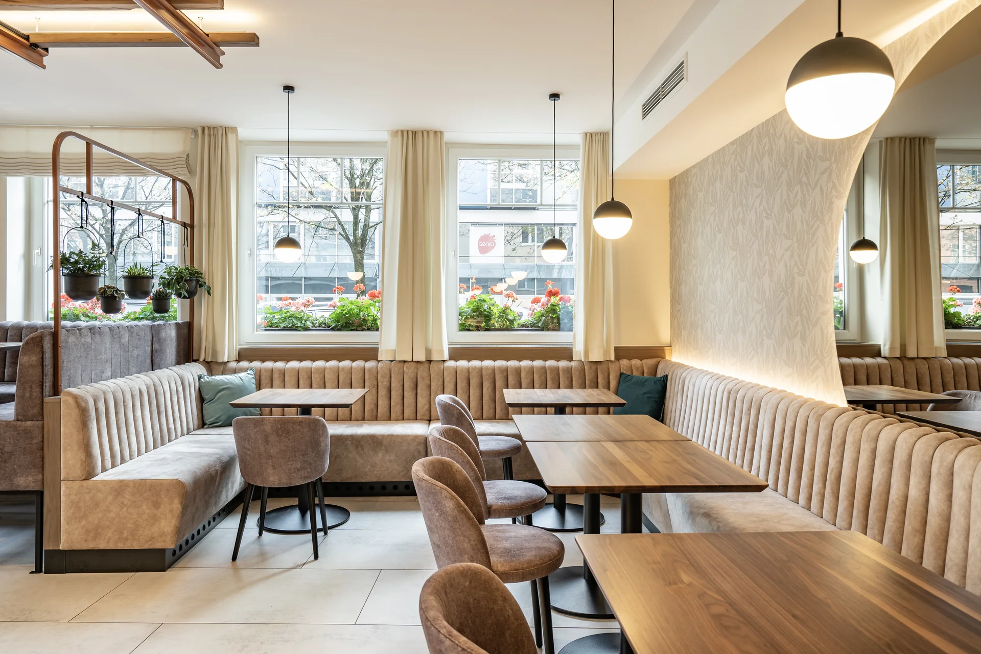 Frühstücksbereich im Hotel Lokomotive Linz mit langer beigefarbener Samtbank, Holztischen, braunen Samtstühlen, hängenden Zimmerpflanzen an einer Kupferstange und Panoramafenstern mit Blick auf rote Geranien.