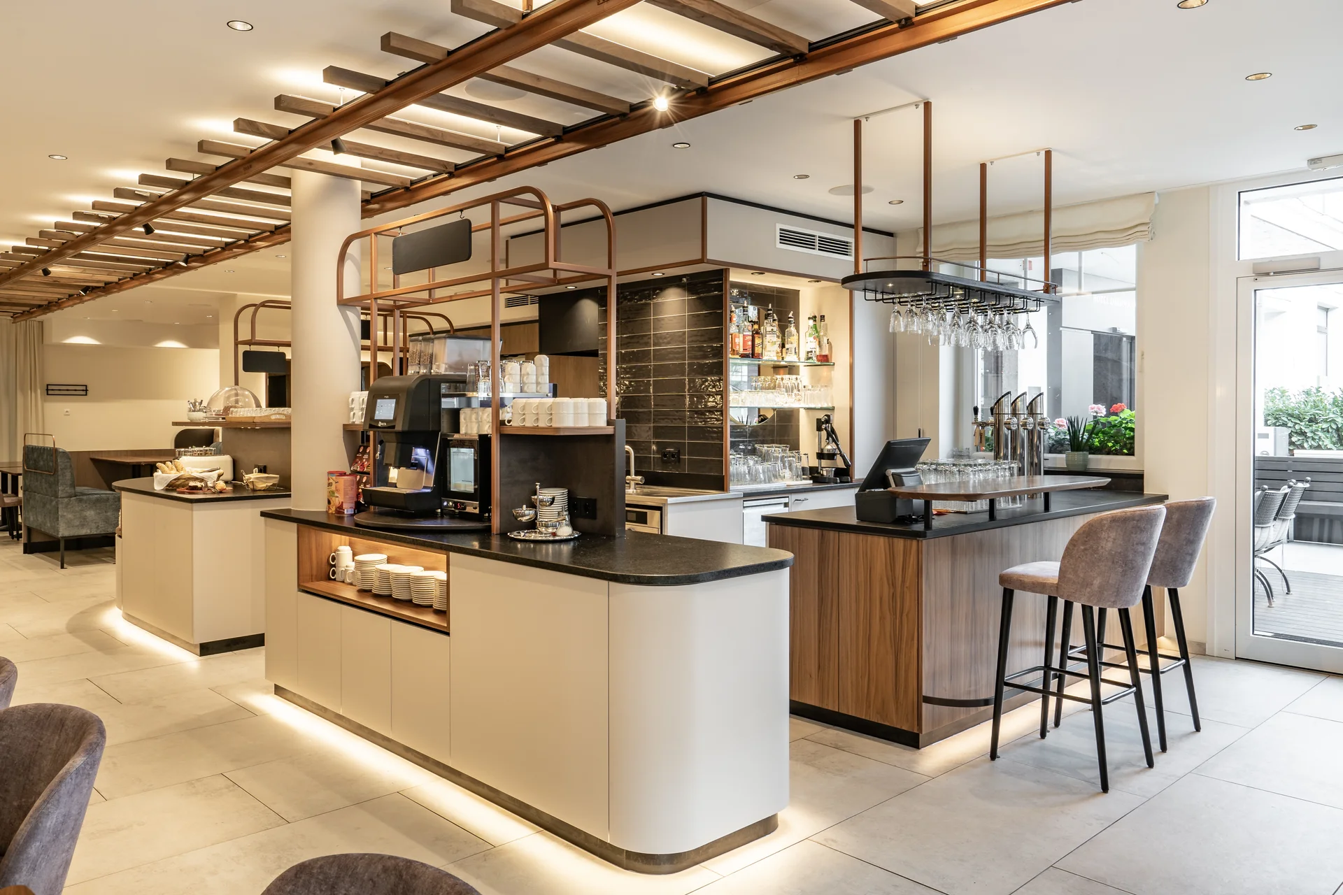 Bar- und Kaffeestation im Hotel Lokomotive Linz mit geschwungenen hellen Tresen, schwarzer Granitarbeitsfläche, Kupfer-Holz-Deckenregal mit hängenden Weingläsern, Kaffeemaschine und Blick auf den Frühstücksbereich.