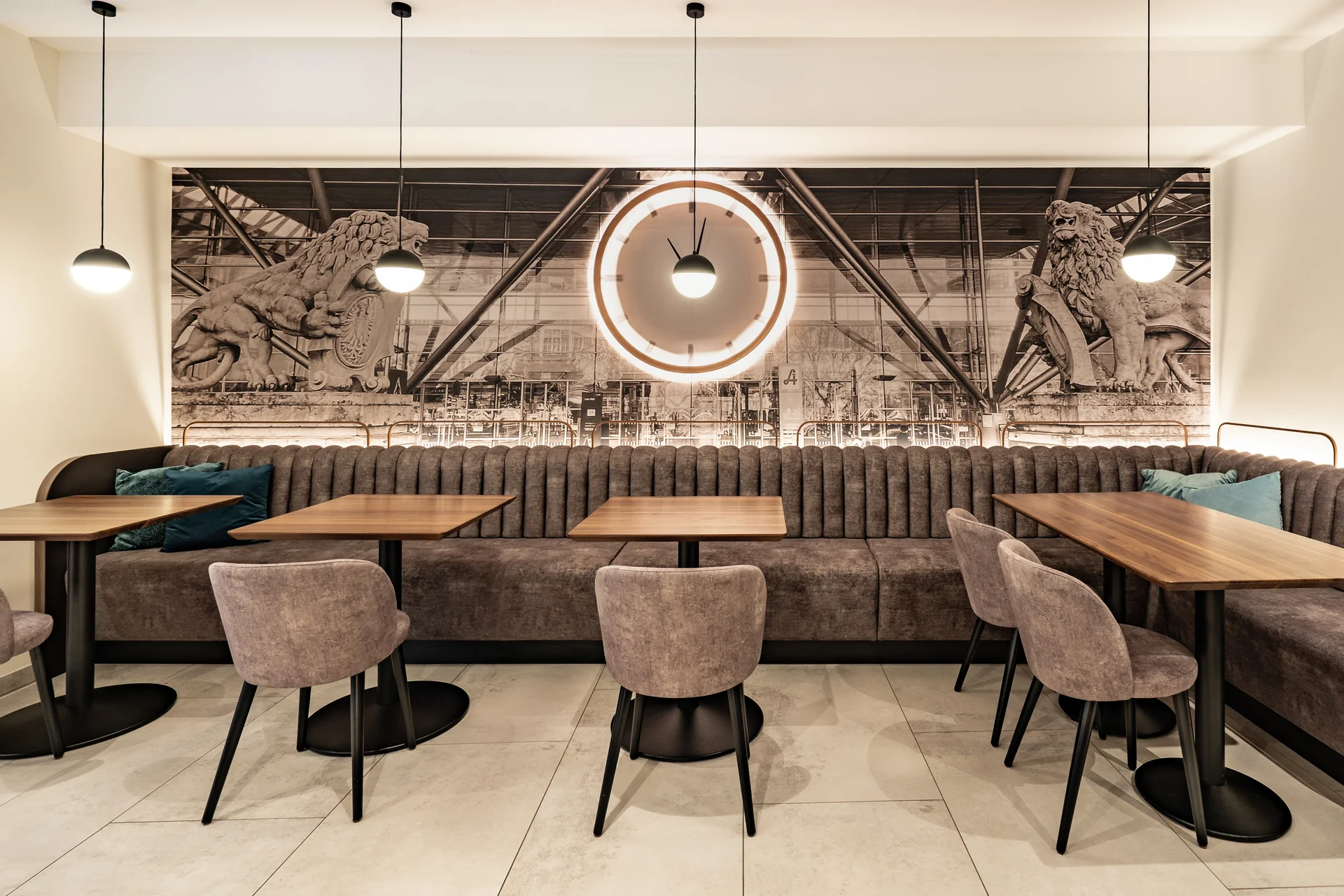 Restaurantbereich im Hotel Lokomotive Linz mit langer grauer Samtbank, Holztischen, grauen Polsterstühlen mit türkisen Kissen und großformatigem Schwarz-Weiß-Wandbild des Linzer Bahnhofs mit Löwenskulpturen und rundem Wandspiegel.