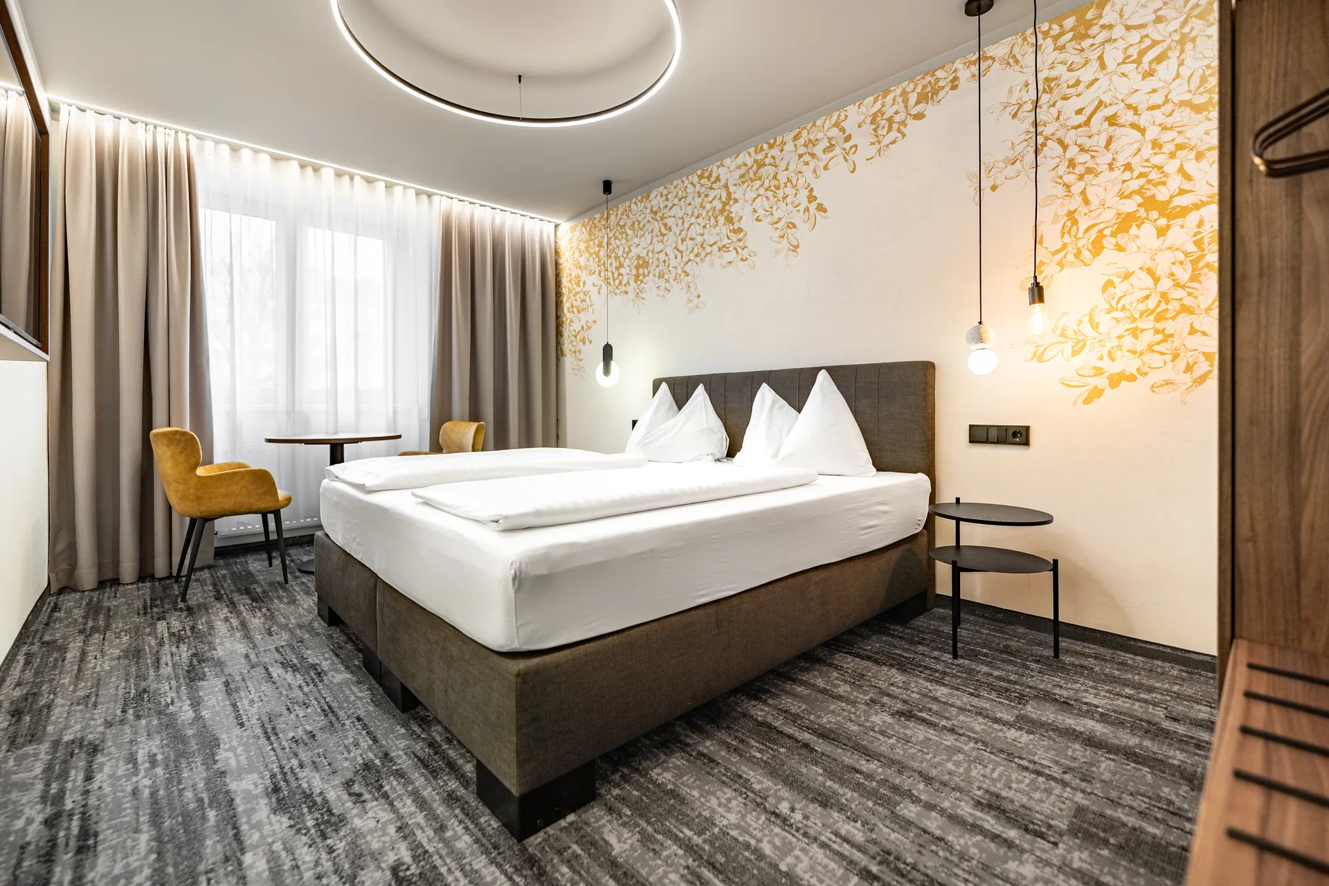 Hotelzimmer im Hotel Lokomotive Linz mit braunem Boxspringbett, weißer Bettwäsche, goldenem Blattmotiv an der Wand, rundem Deckenlichtring, senfgelben Sesseln am Fenster und gemustertem Teppich.