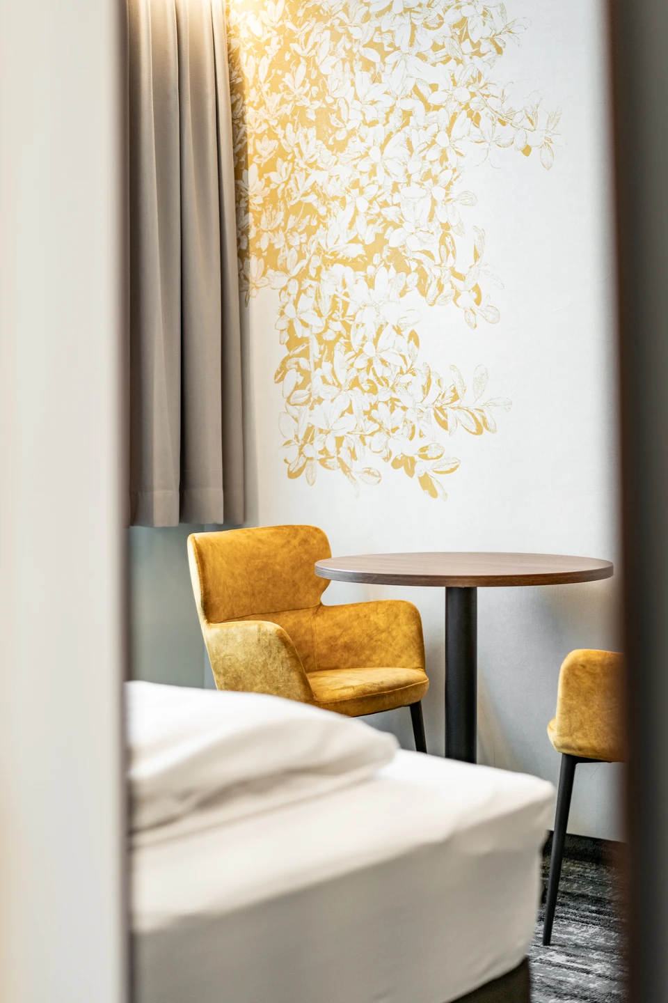 Hotelzimmer im Hotel Lokomotive Linz mit zwei senfgelben Samtsesseln und rundem Holztisch vor einer Wand mit goldenem Blatt- und Blütenmotiv, im Vordergrund das weiße Bett.