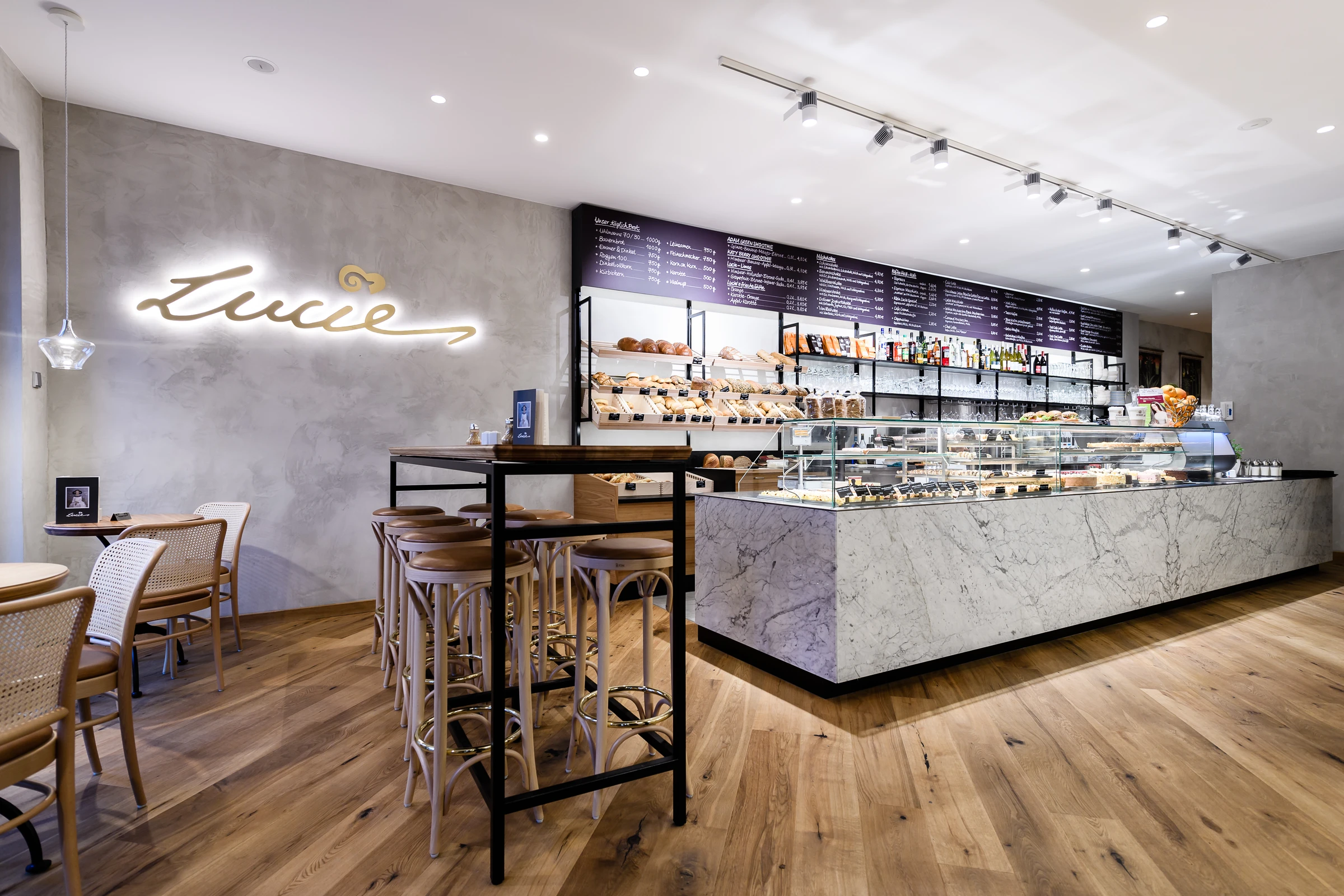 Innenbereich des Café Lucie mit marmorverkleideter Verkaufstheke mit Backwaren und Kuchen, Holzbarhockern an einem Stehtisch, beleuchteter Lucie-Schriftzug an der Betonwand und Wiener Korbgeflecht-Stühlen im Hintergrund.