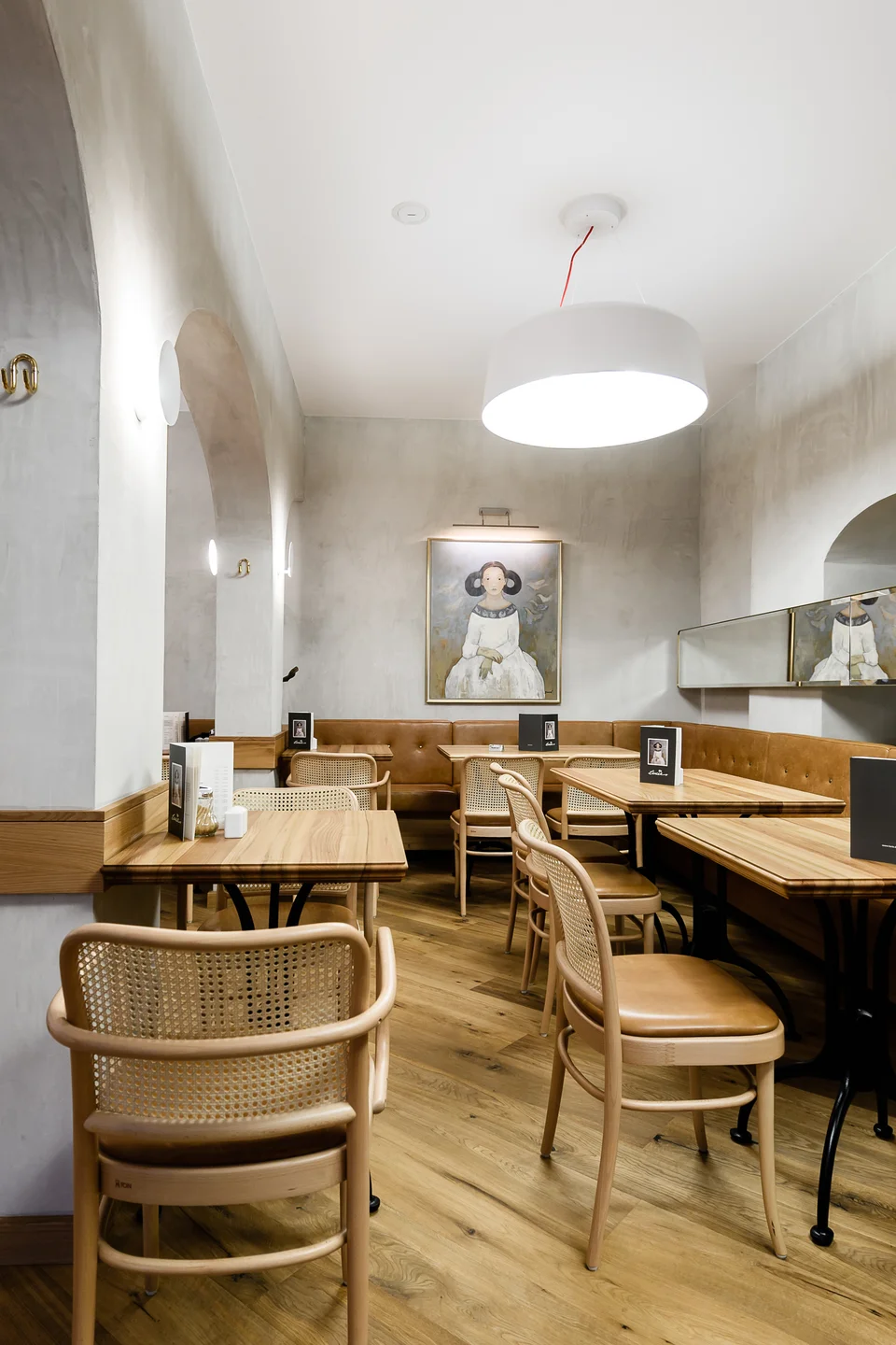 Cafébereich des Café Lucie Wien mit historischen Gewölbebögen, cognacfarbener Lederbank, Wiener Korbgeflecht-Stühlen, Holztischen und einem gerahmten figurativen Gemälde an der Wand.