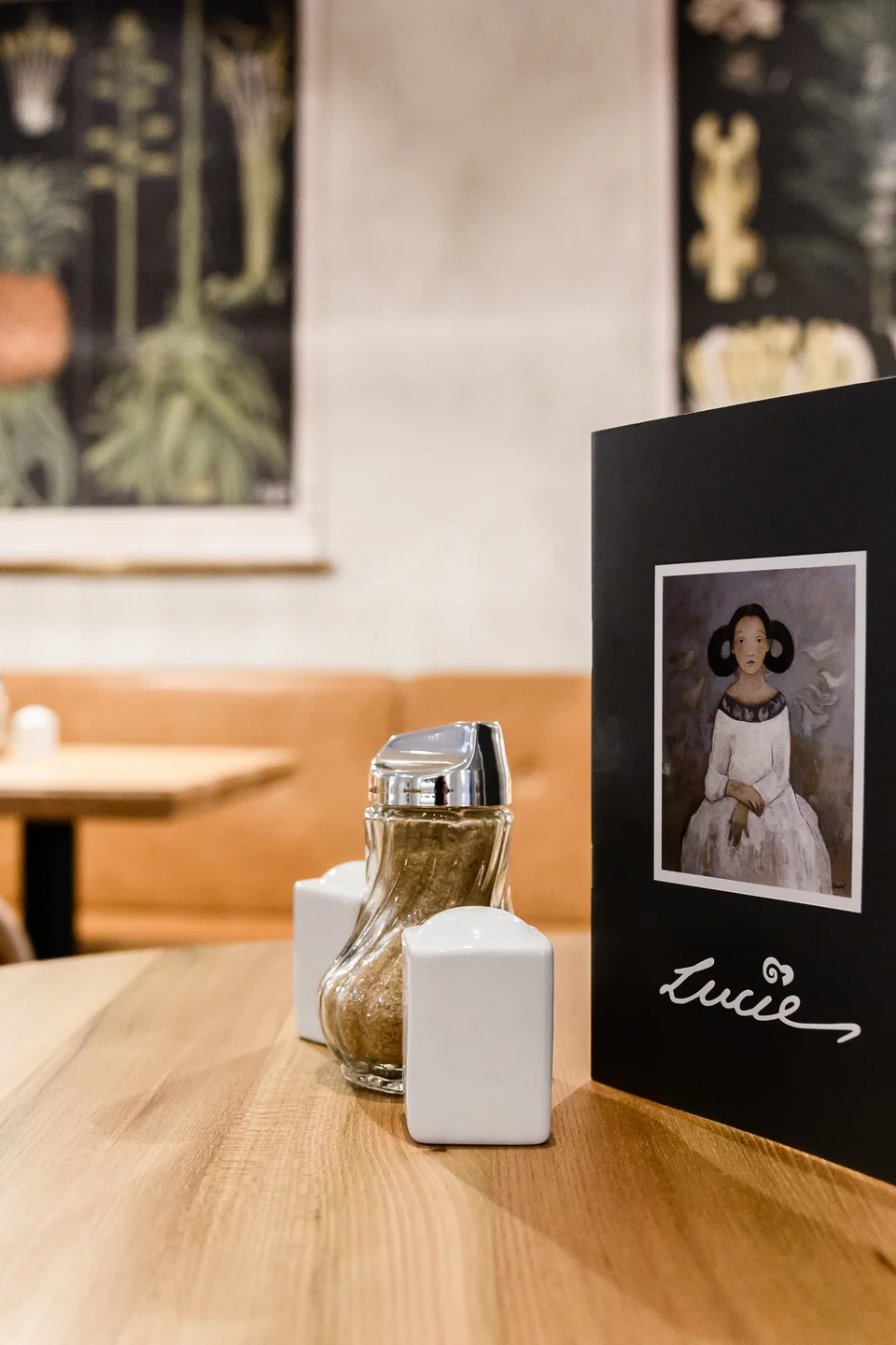 Tischdetail im Café Lucie Wien mit schwarzer Speisekarte mit Lucie-Schriftzug, Salz- und Pfefferstreuer auf hellem Holztisch, im Hintergrund cognacfarbene Lederbank und botanische Wandbilder.