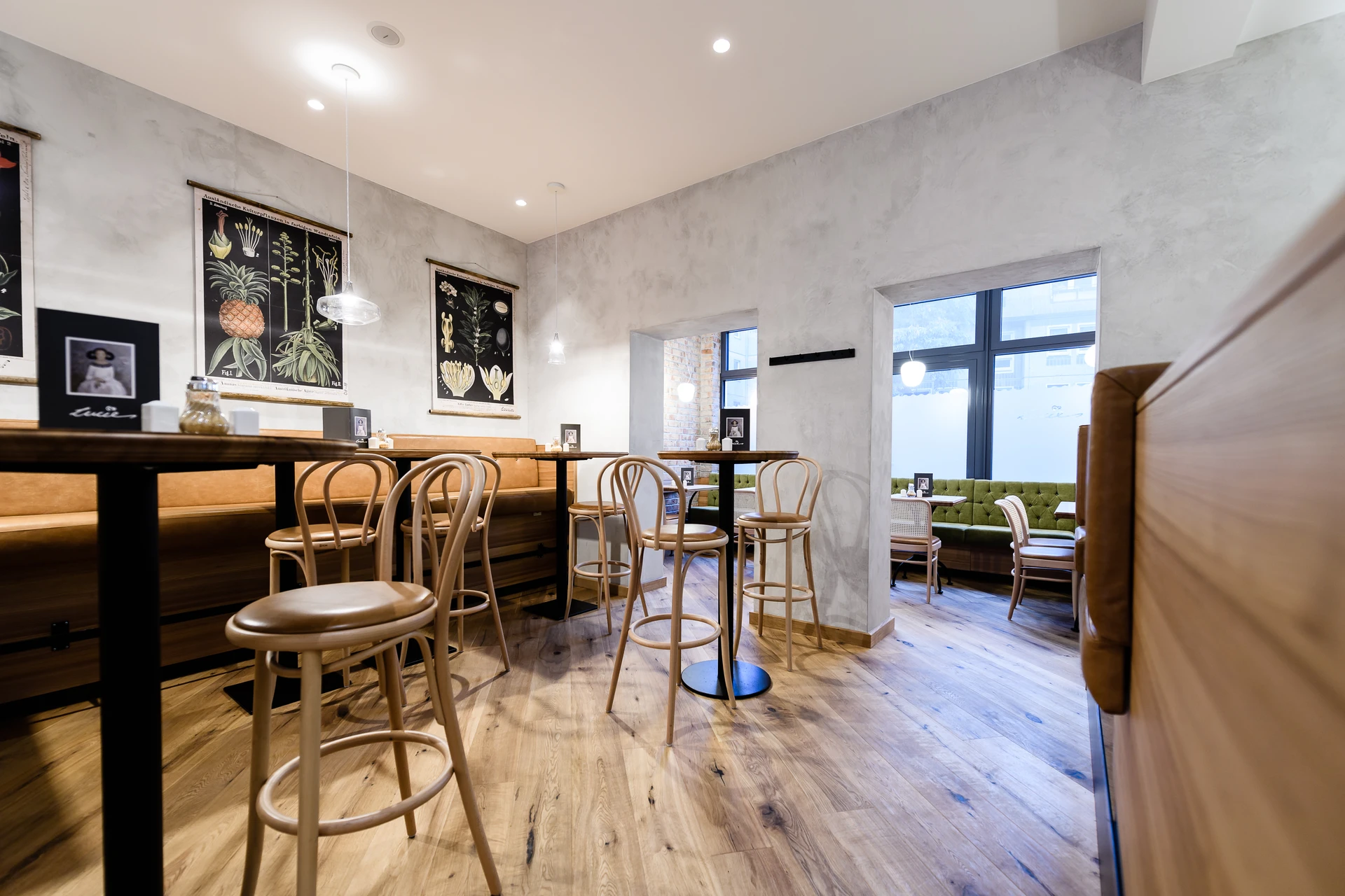 Cafébereich des Café Lucie Wien mit Holzbarhockern an langen Stehtischen, cognacfarbener Lederbank, botanischen Wandbildern auf Betonputzwand, Holzparkett und Blick in den hinteren Gastraum mit grüner Samtbank.