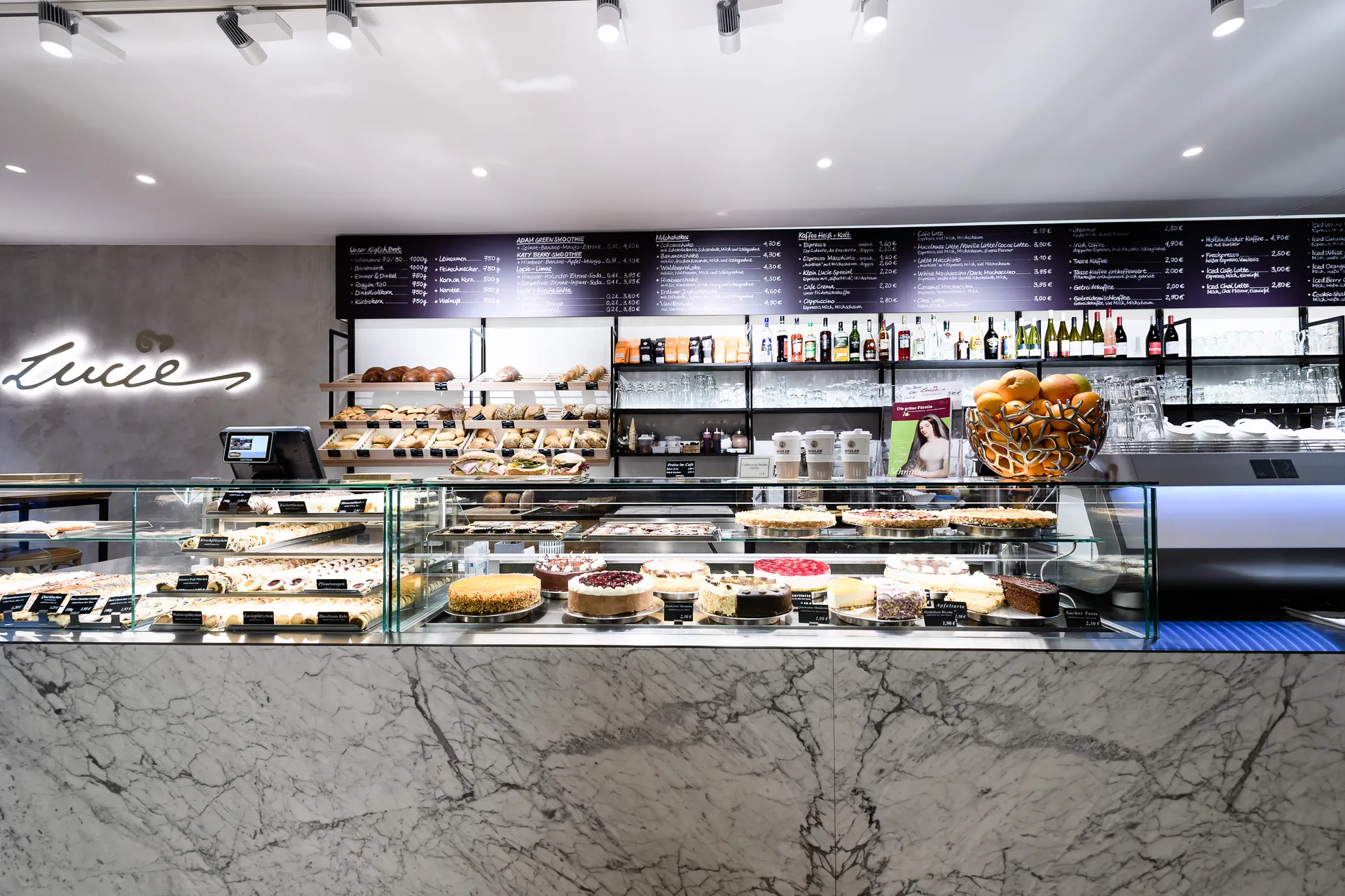 Verkaufstheke im Café Lucie Wien mit marmorverkleideter Front, Kuchentheke mit verschiedenen Torten, Backwarenregal, dunkler Tafelkarte mit Speiseangebot und beleuchtetem Lucie-Schriftzug an der Wand.