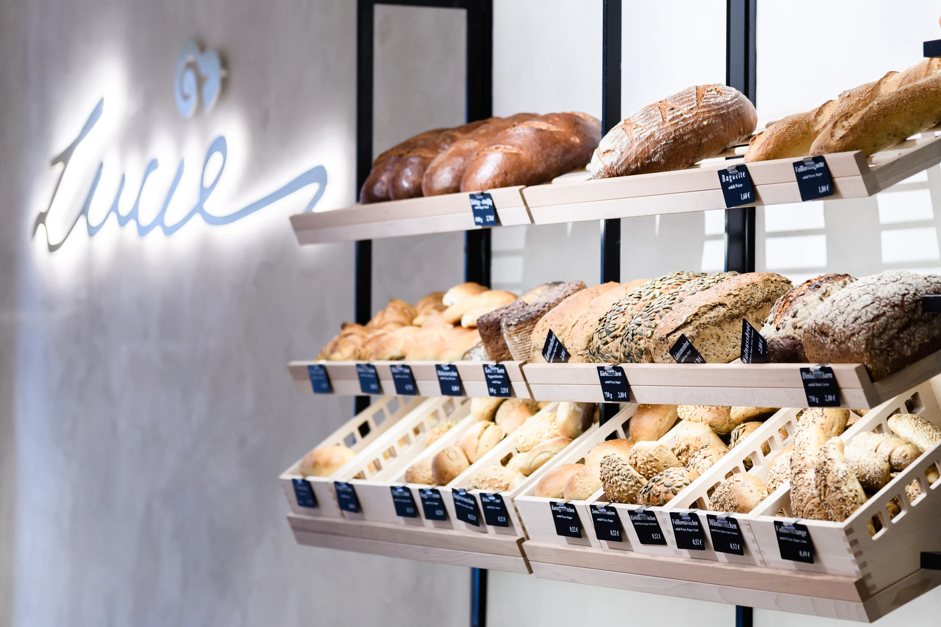 Backwarenregal im Café Lucie Wien mit verschiedenen Broten, Baguettes und Semmeln in weißen Holzkörben mit Preisschildern, links im Hintergrund der beleuchtete Lucie-Schriftzug an der Betonputzwand.