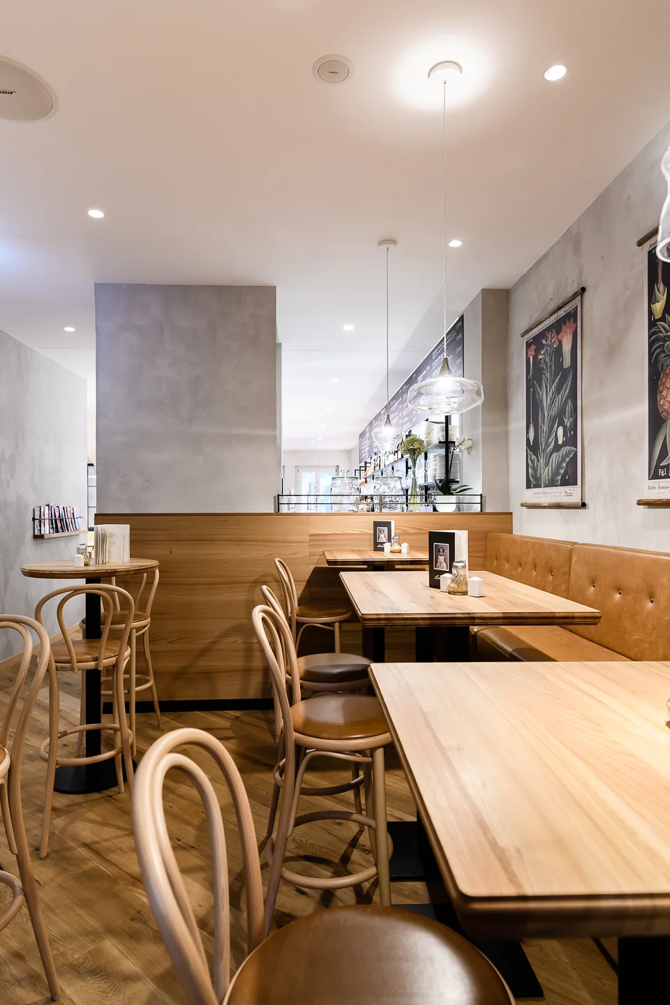 Gastraum im Café Lucie Wien mit cognacfarbener Lederbank, Holztischen, Wiener Korbgeflecht-Stühlen, Glaspendelleuchten und botanischen Wandbildern auf Betonputzwand.