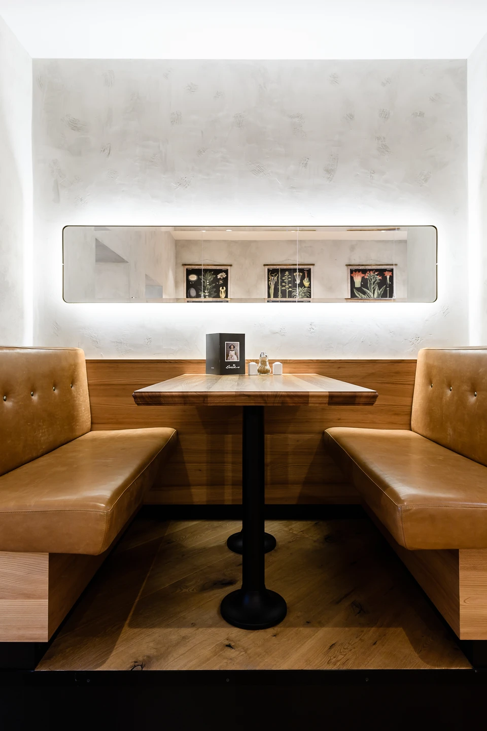 Gemütliche Dinerbänke im Café Lucie Wien mit cognacfarbener Lederbank, Holzverkleidung, beleuchtetem Wandspiegel in dem sich botanische Bildern spiegeln.