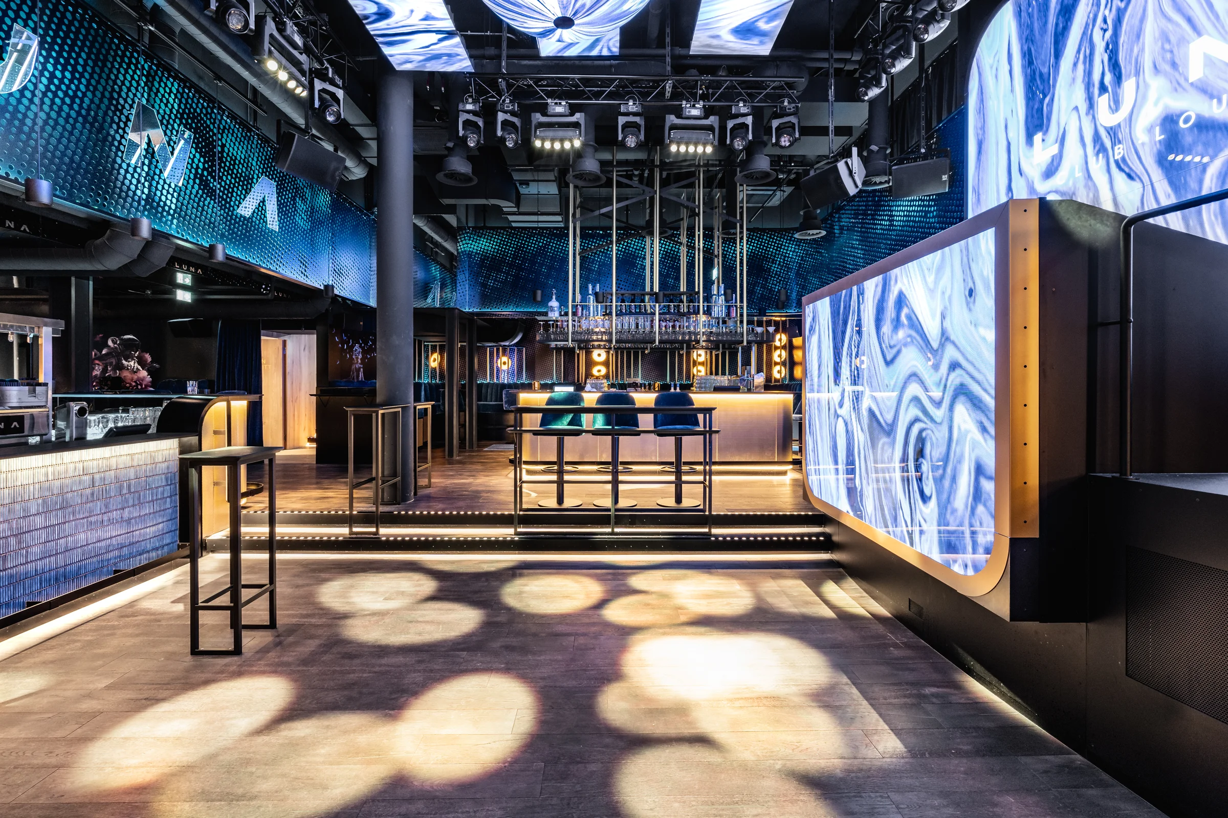 Innenbereich des Club Luna Innsbruck mit Tanzfläche, hinterleuchtetem Bartresen mit blauen Barhockern, großem LED-Screen mit abstraktem Muster, blauer LED-Deckeninstallation und Bühnenbeleuchtung.