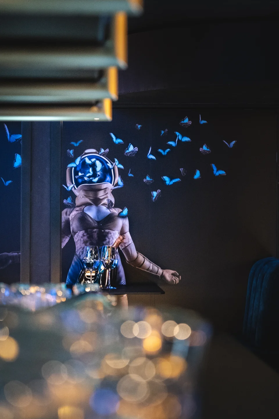 Tapete mit Animation im Club Luna Innsbruck mit einer futuristischen Figur in Rüstung umgeben von blauen Schmetterlingen, im Vordergrund unscharf Weingläser und Bokeh-Lichteffekte.