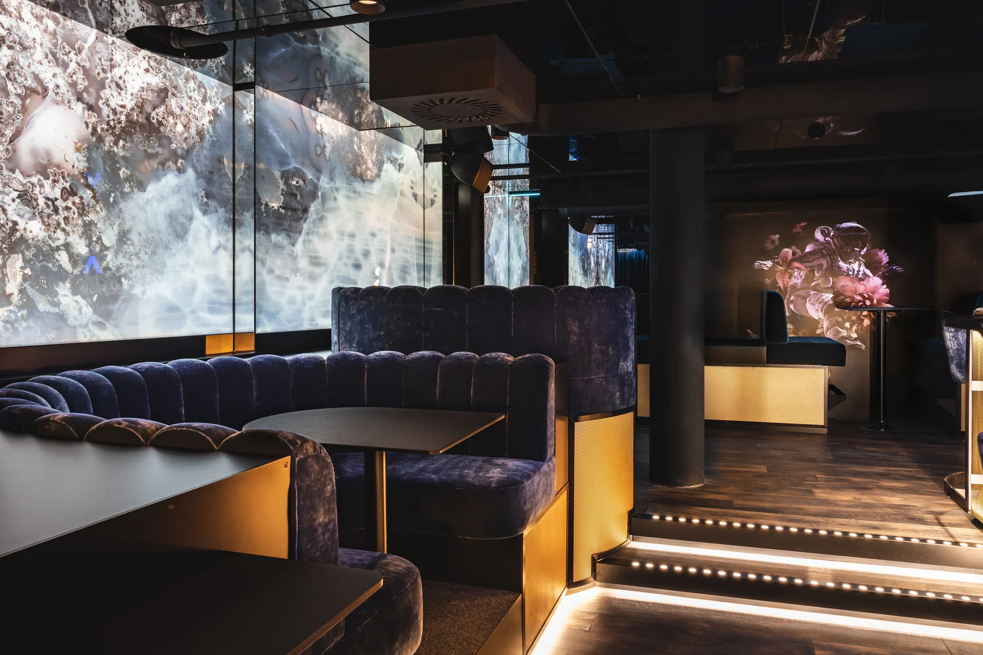 VIP-Loungebereich im Club Luna Innsbruck mit dunkelblauem Samtrundsofas mit Goldverkleidung, schwarzen Tischen, hinterleuchtetem LED-Screen mit abstraktem Muster und beleuchteten Treppenstufen.