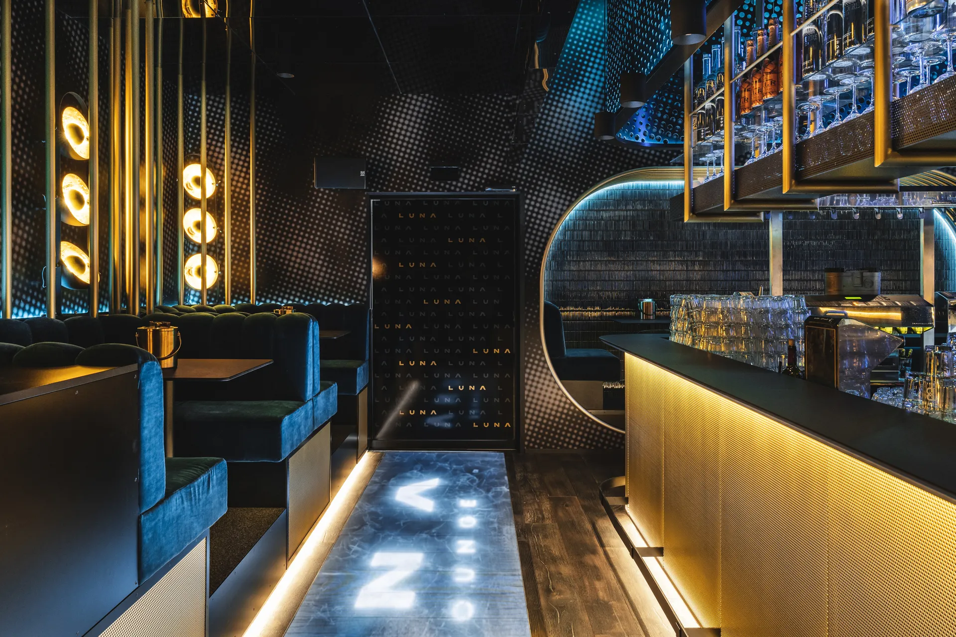 Bar- und Loungebereich im Club Luna Innsbruck mit goldenem Bartresen, dunkelgrünen Chesterfield-Samtsofas, goldenen Wandleuchten, am Boden leuchtendes LUNA-Logo und hängendem Weinglasregal.