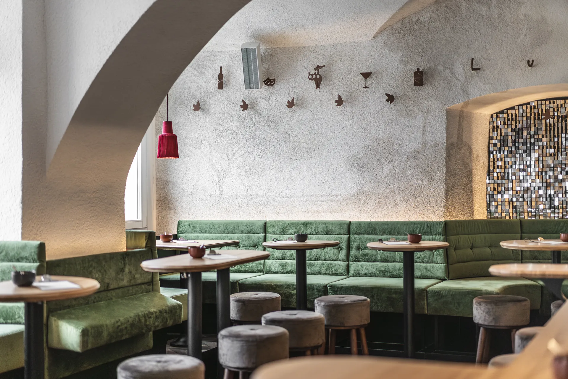 Gastraum des Lust in Bregenz mit grüner Samtbank, runden Holztischen und verspielten Metall-Vogelskulpturen an der Gewölbewand neben einem goldenen Mosaikbogen.