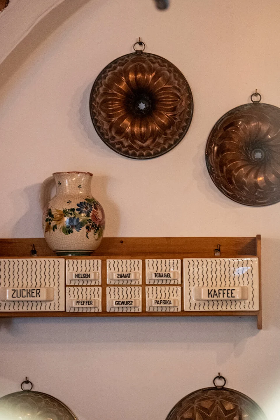 Vintage Gewürzregal mit beschrifteten Keramikschubladen und bemaltem Krug im Gasthaus Maderthaner Weistrach, flankiert von dekorativen Kupfer-Backformen an der Wand.