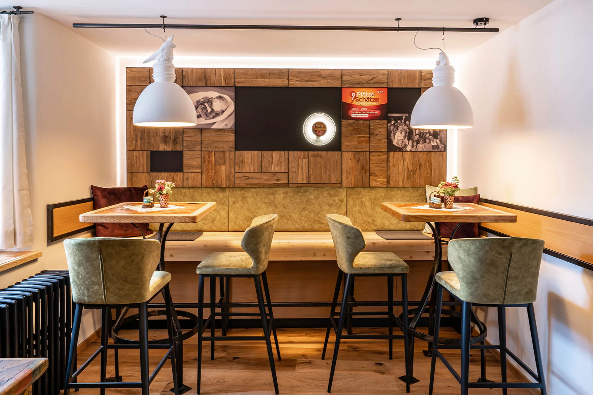 Moderner Barbereich im Gasthaus Maderthaner Weistrach mit grünen Barhockern, Naturholztischen, Altholzwand mit Auszeichnungen und weißen Hängelampen.