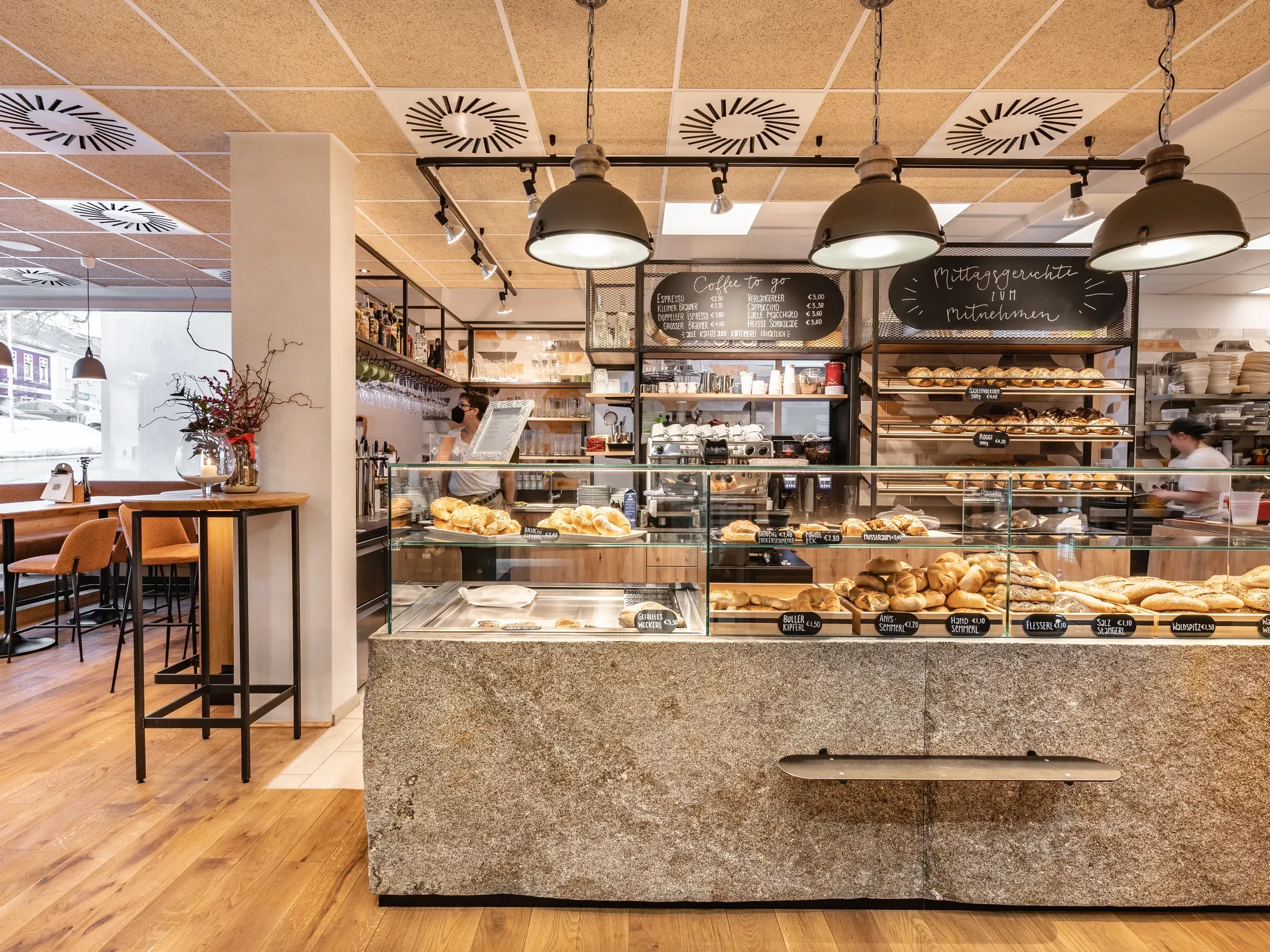 Verkaufstheke der Bäckerei Manuell in Oberneukirchen mit reichhaltigem Brotsortiment, Kreidetafeln für Coffee-to-go und Mittagsgerichte sowie Industriependelleuchten über dem Granittresen.