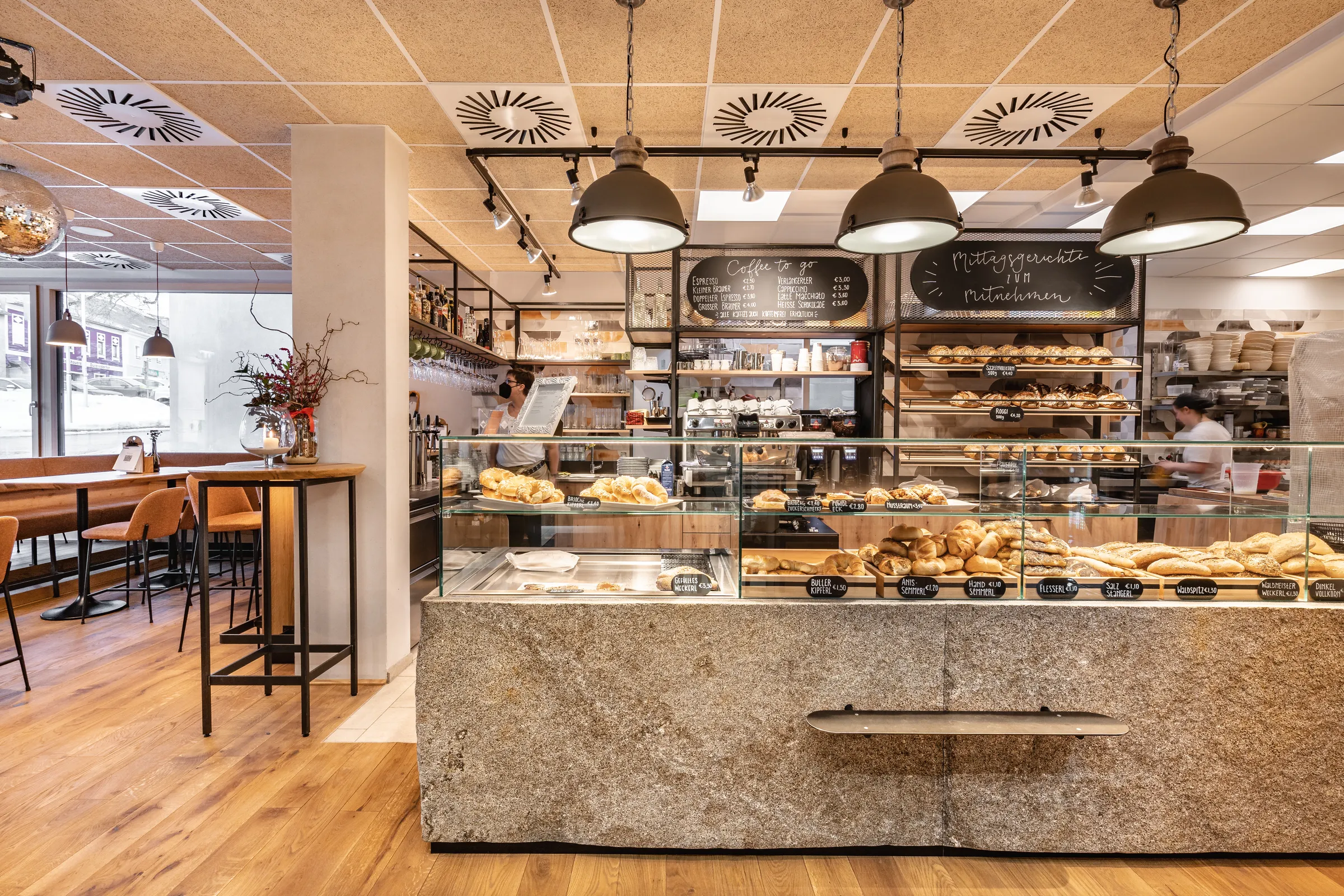 Verkaufstheke der Bäckerei Manuell in Oberneukirchen mit reichhaltigem Brotsortiment, Kreidetafeln für Coffee-to-go und Mittagsgerichte sowie Industriependelleuchten über dem Granittresen.