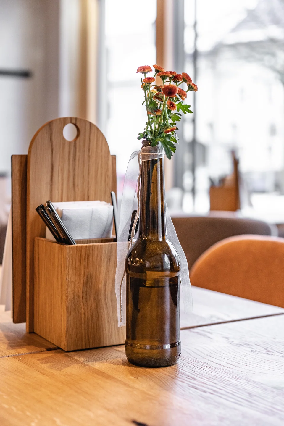 Tischdetail der Bäckerei Manuell in Oberneukirchen mit einer Glasflasche als Vase mit orangefarbenen Blumen und einem Holzständer für Servietten und Besteck auf hellem Eichenholztisch.