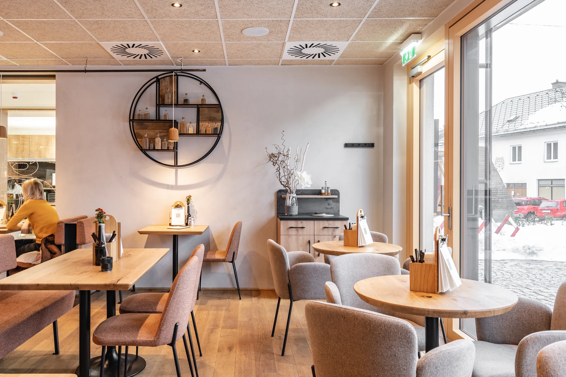 Heller Cafébereich der Bäckerei Manuell in Oberneukirchen mit Eichenholztischen, gemischten Sesseln und rundem Wandregal, mit Blick durch große Panoramafenster auf den verschneiten Marktplatz.