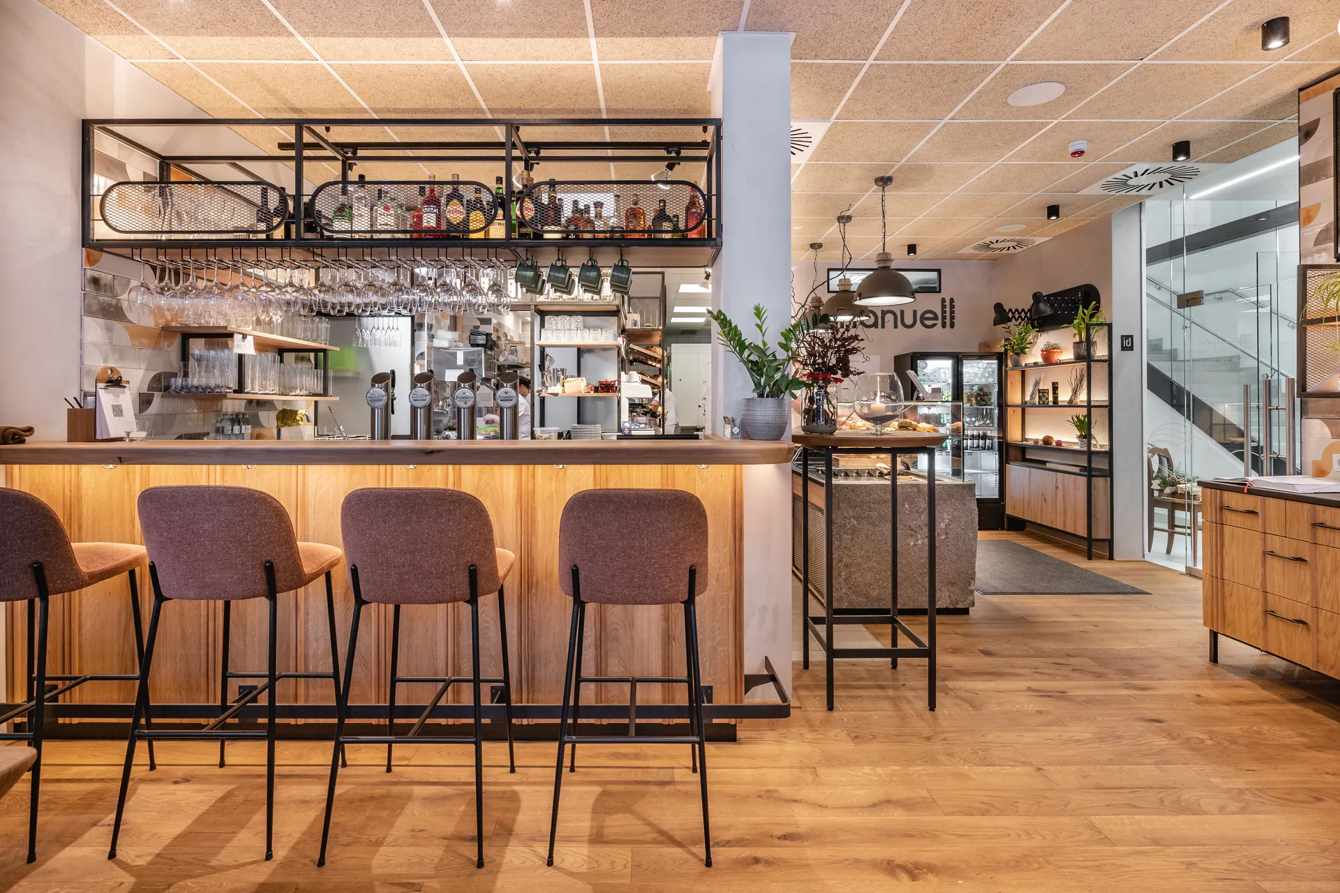 Barbereich der Bäckerei Manuell in Oberneukirchen mit hellem Holztresen, Barhockern und hängendem Glasregal über der Theke, im Hintergrund der Logoschriftzug und die Verkaufstheke.