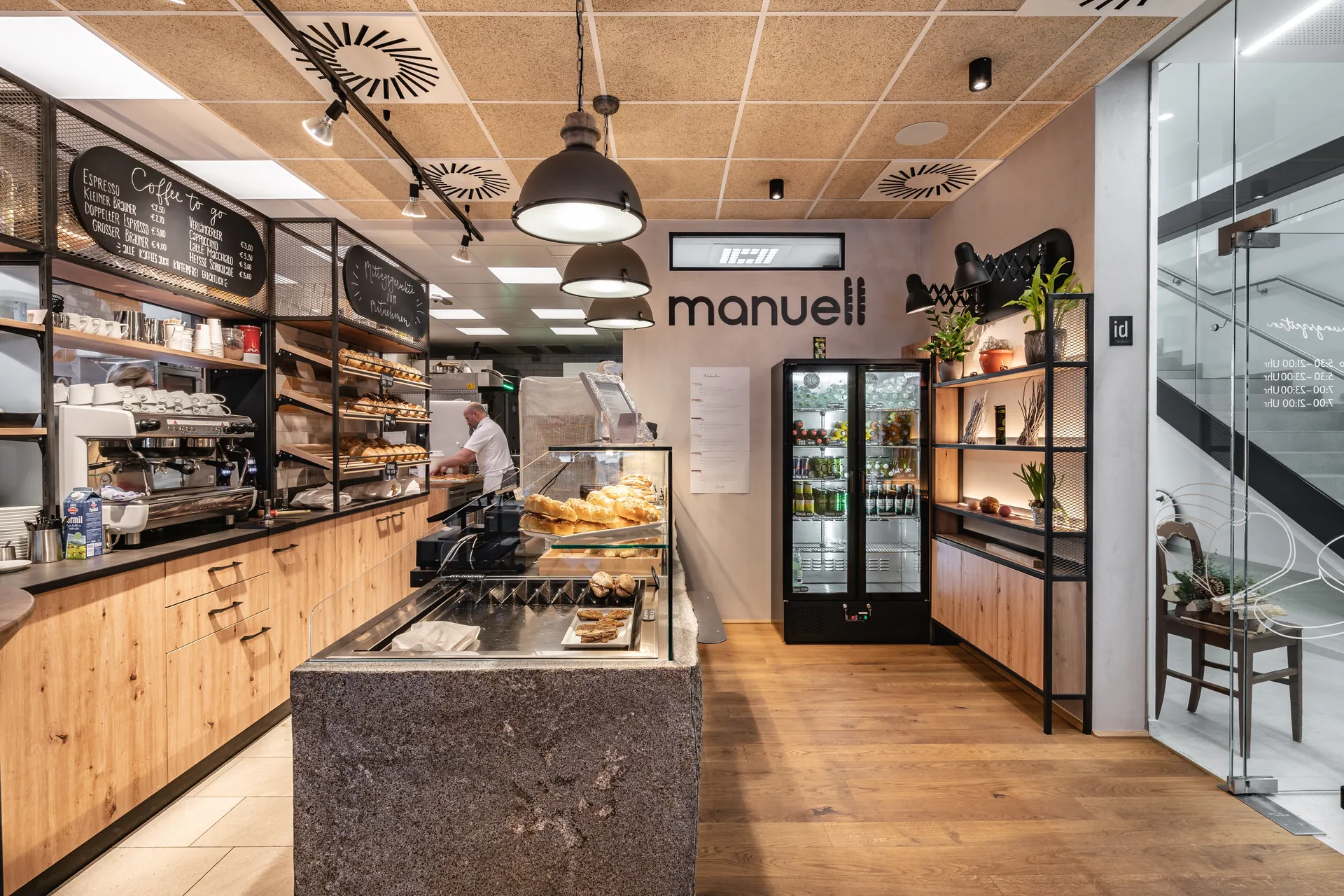 Verkaufsbereich der Bäckerei Manuell in Oberneukirchen mit Backwarentheke, Logoschriftzug, Getränkekühlschrank und beleuchtetem Regal neben dem verglasten Treppenaufgang.