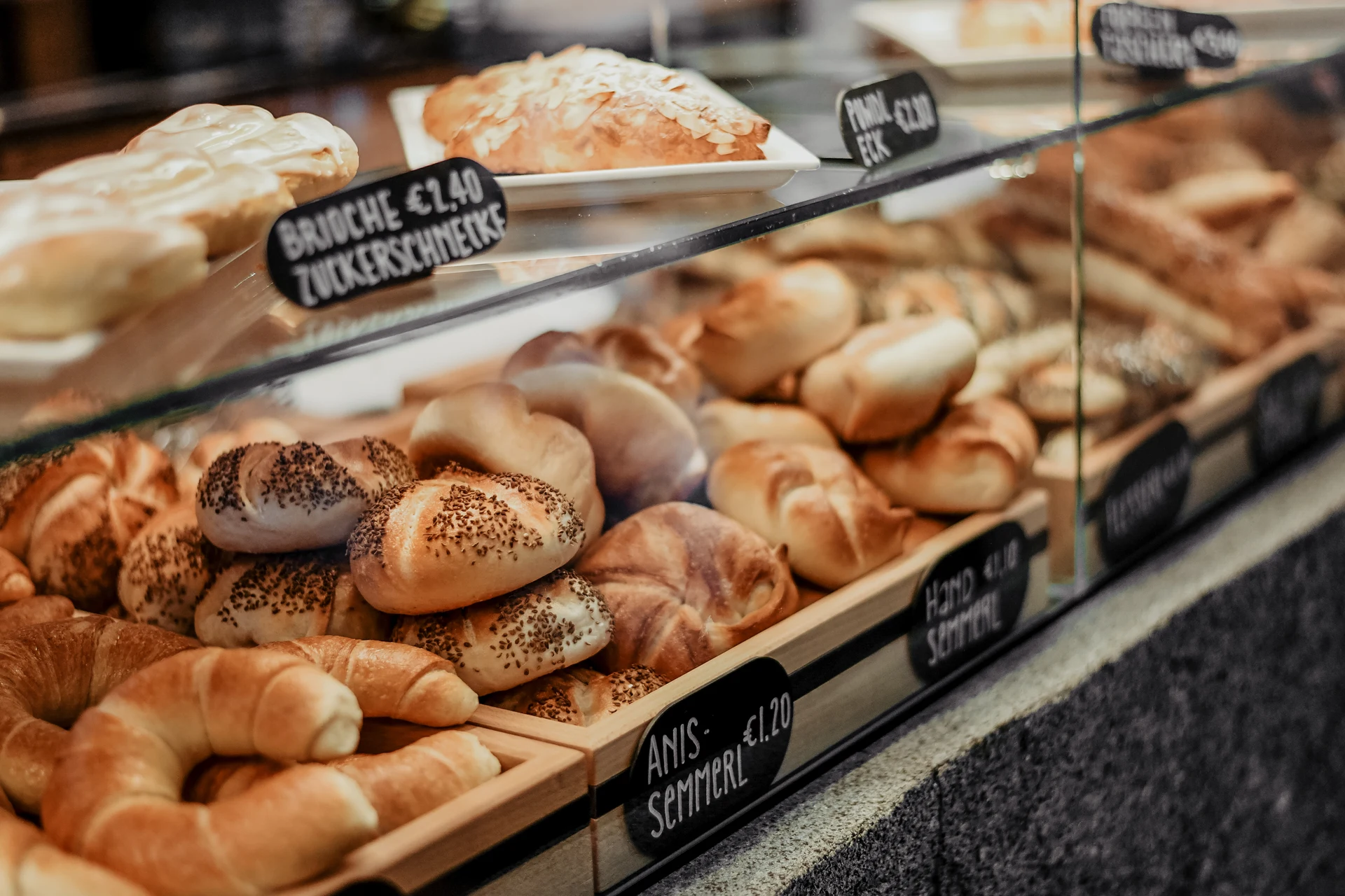 Handgeschriebene Kreidetafel mit dem Coffee-to-go-Angebot der Bäckerei Manuell in Oberneukirchen, eingerahmt in ein schwarzes Metallgitterregal unter einer Industriependelleuchte.