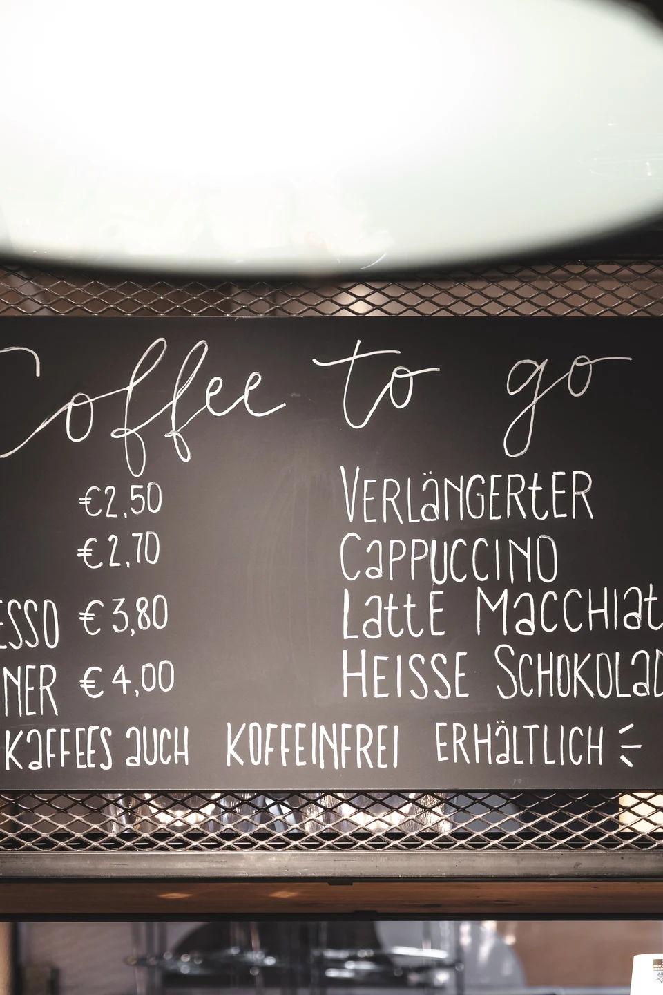 Handgeschriebene Kreidetafel mit dem Coffee-to-go-Angebot der Bäckerei Manuell in Oberneukirchen, eingerahmt in ein schwarzes Metallgitterregal unter einer Industriependelleuchte.