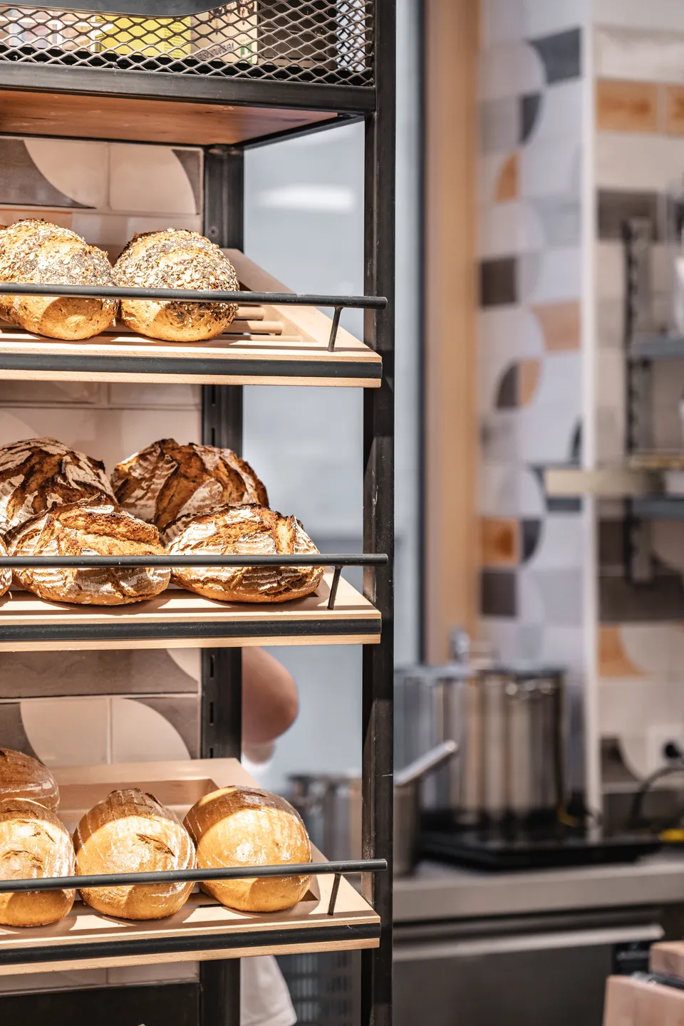 Brotregal der Bäckerei Manuell in Oberneukirchen mit frisch gebackenen Sauerteigbroten und Semmerln auf mehreren Etagen, im Hintergrund die Backstube mit gemusterter Fliesenwand.