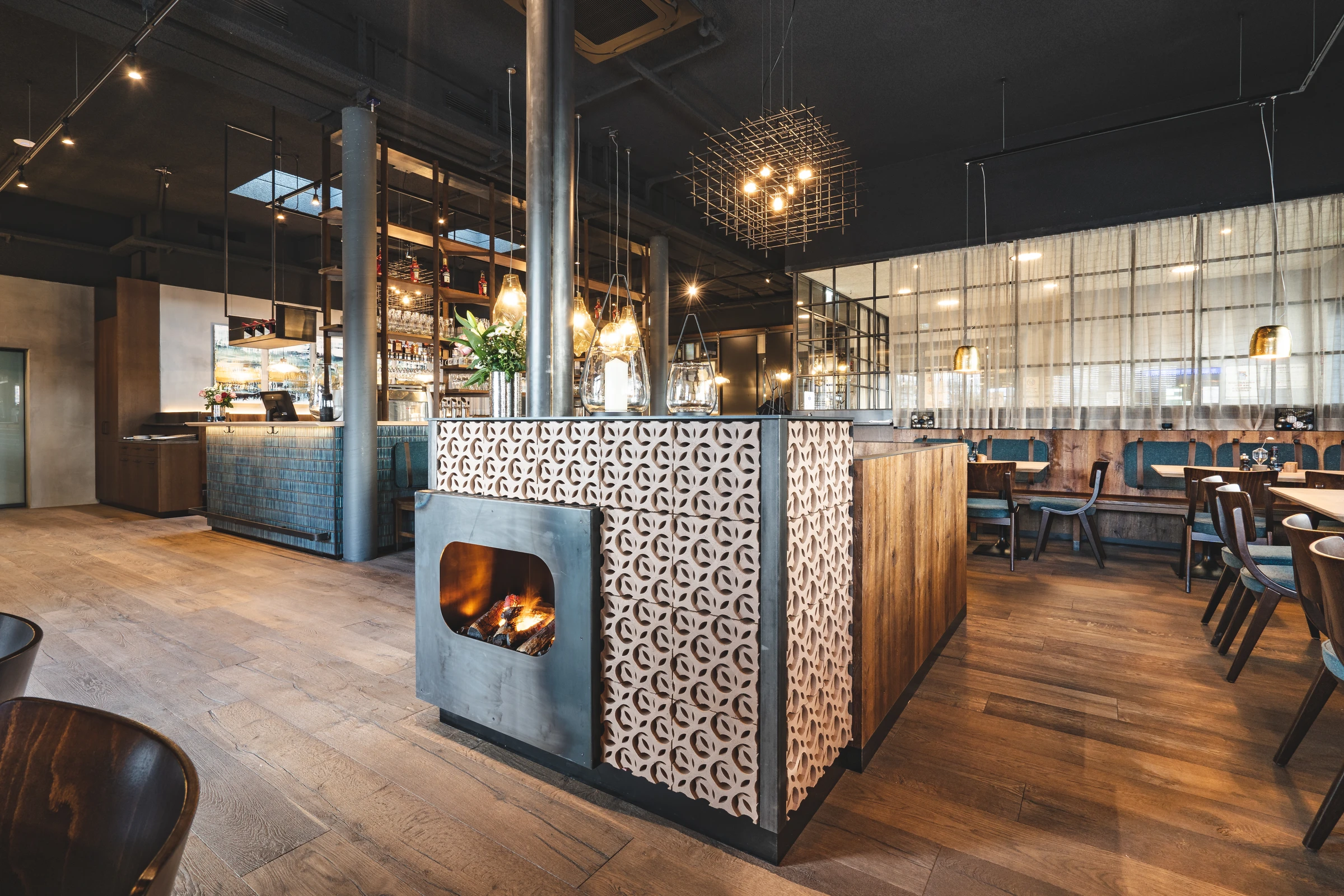 Gesamtansicht des Restaurants Miraculix in Natternbach mit zentralem Kamin-Raumteiler aus Betonkacheln, Holzparkett, freistehendem Bartresen und blauer Sitzbank im Hintergrund.