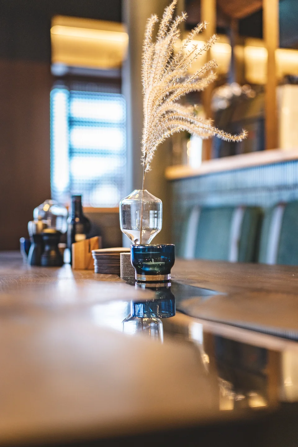 Tischdetail im Restaurant Miraculix in Natternbach mit einer Glasvase mit Pampasgras und blauem Teelicht auf polierter Tischfläche, im Hintergrund die blaue Sitzbank und ein beleuchtetes Regal.