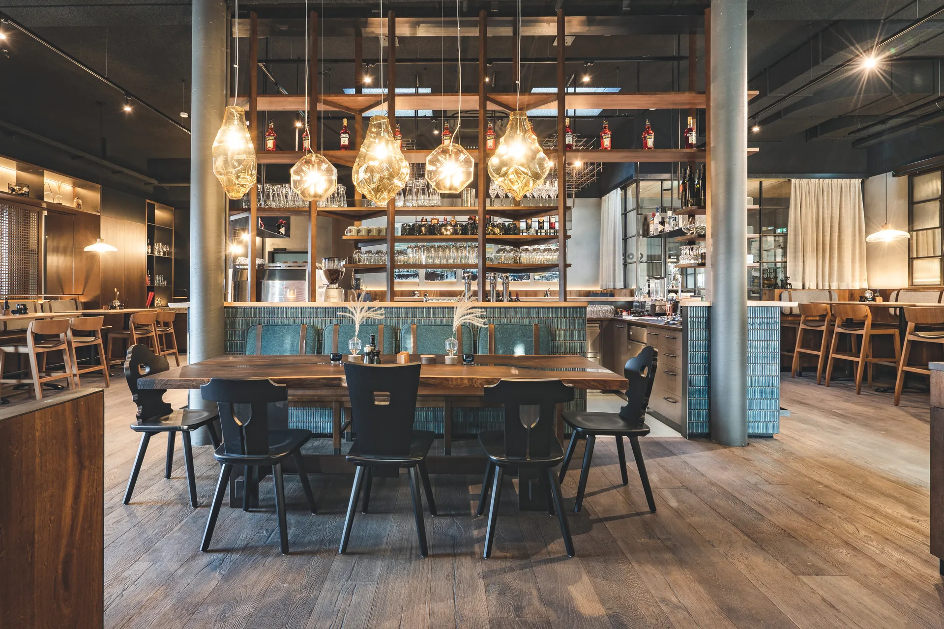 Gastraum des Restaurants Miraculix in Natternbach mit massivem Holztisch, schwarzen Trachtstühlen und einer imposanten Barwand mit Holzregal, türkisen Kacheln und großen Glaspendelleuchten.