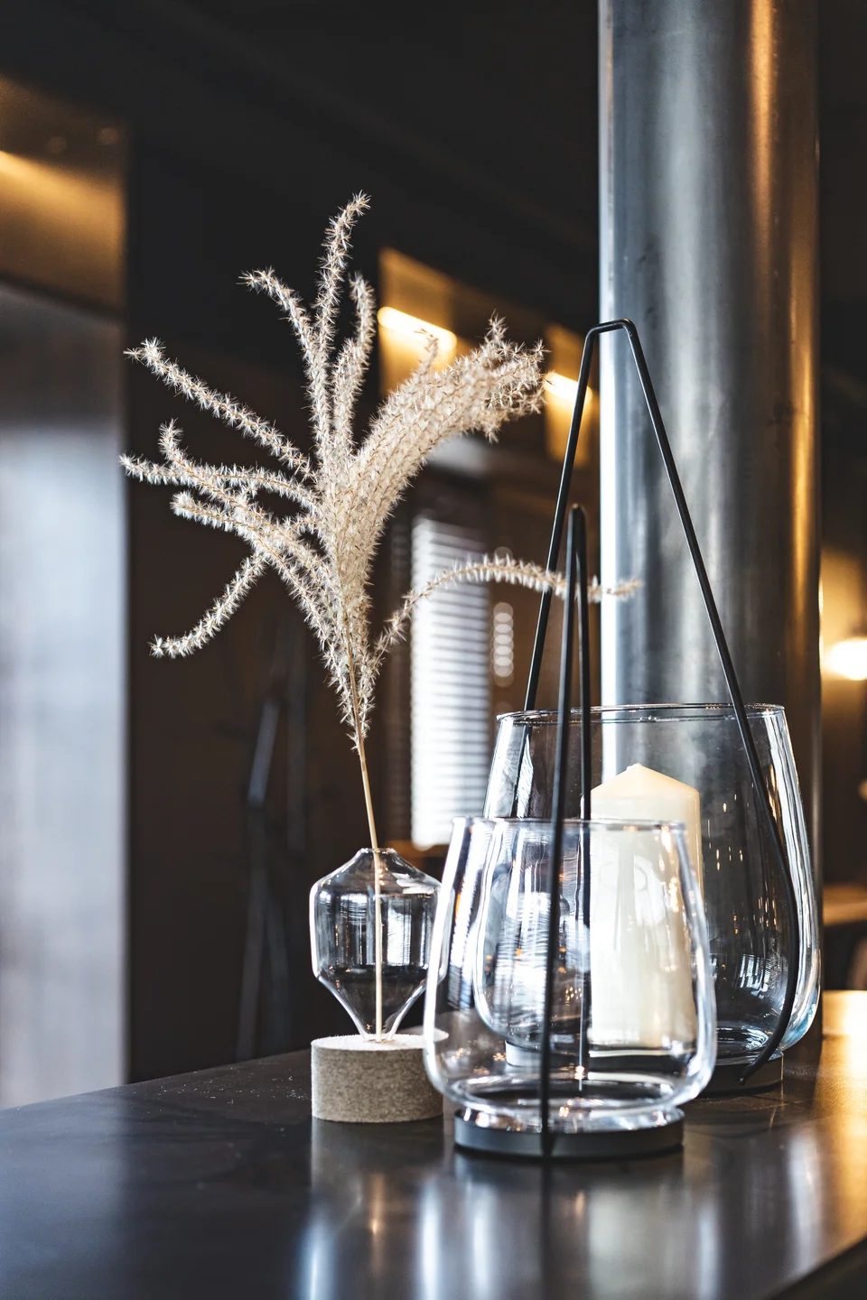 Tischdekoration im Restaurant Miraculix in Natternbach mit Pampasgras in einer Glasvase und einem Windlicht im schwarzen Metallgestell auf dunkler Tresenfläche.