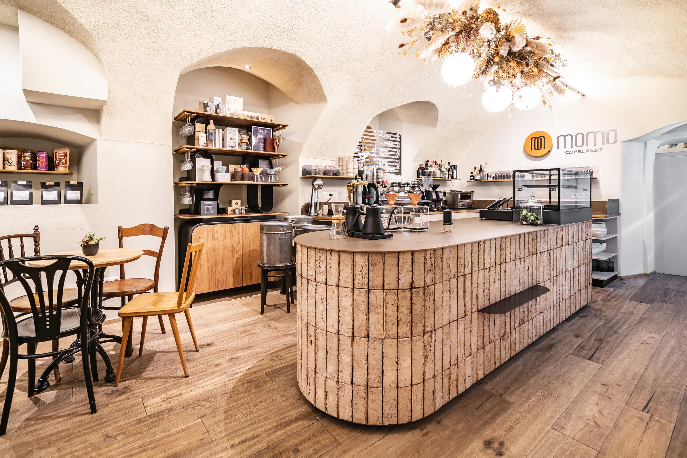 Gesamtansicht des Café Momo Coffeeart in Feldkirch mit gerundetem Terrakottafliesentresen, historischem Gewölbe, Trockenblumen-Kronleuchter und Logoschriftzug an der Wand.