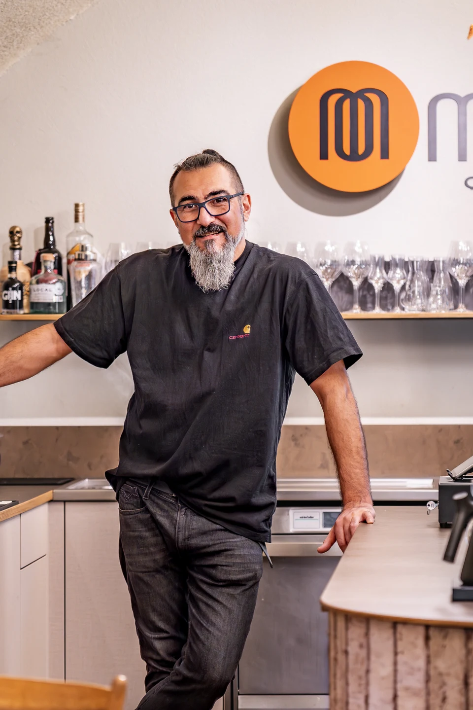 Inhaber des Café Momo Coffeeart in Feldkirch lehnt lässig in seiner Bar vor dem Momo-Logoschriftzug zwischen Espressomaschine und Terrakottafliesentresen.