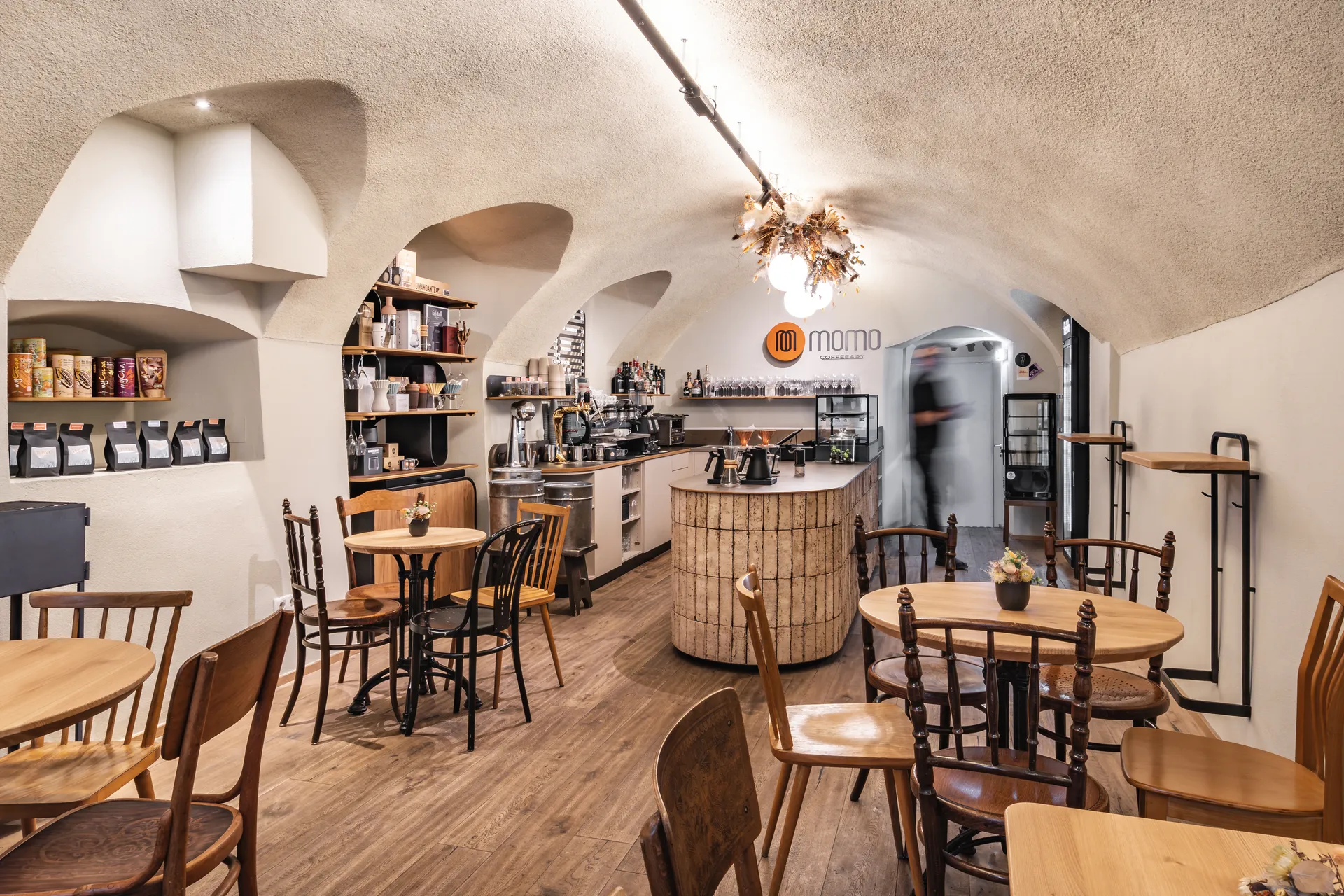 Gesamtansicht des Café Momo Coffeeart in Feldkirch mit historischem Gewölbe, gemischten Holztischen und Thonet-Sesseln, rundem Terrakottafliesentresen und Logoschriftzug an der Wand.