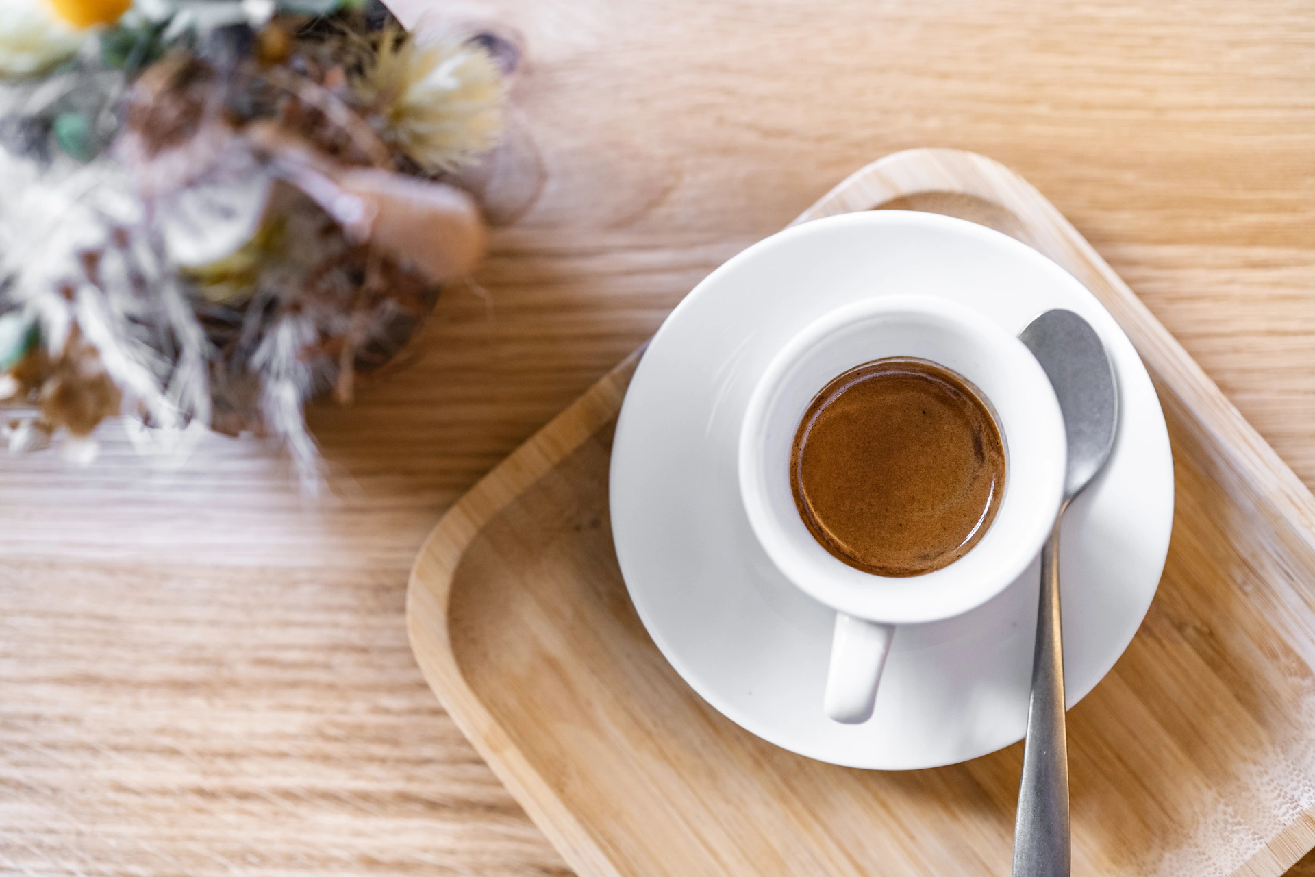 Vogelperspektive auf eine frisch zubereitete Espressotasse mit Crema auf einem Holztablett, daneben ein Trockenblumenstrauß im Café Momo Coffeeart in Feldkirch.
