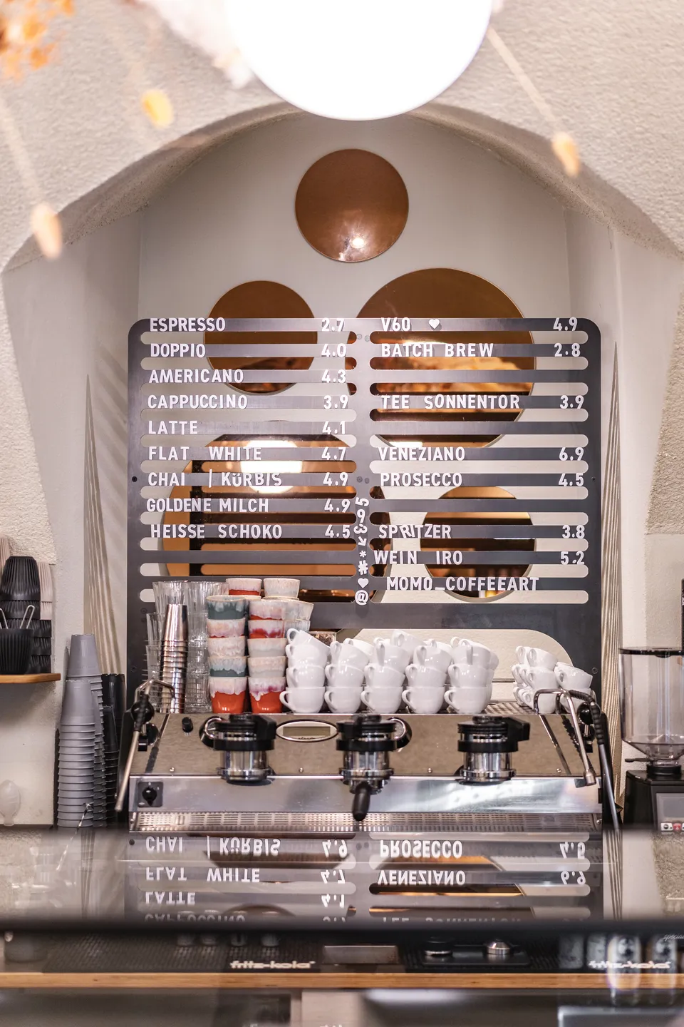 Metallgetränkekarte mit dem vollständigen Kaffeeangebot des Momo Coffeeart in Feldkirch, davor eine La Marzocco Espressomaschine mit bunten Keramik- und weißen Porzellantassen im historischen Gewölbebogen.