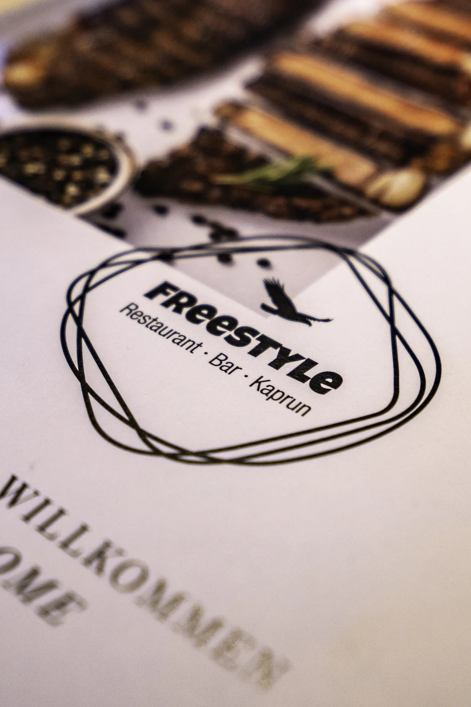 Aufgeschlagene Speisekarte des Restaurant Freestyle in Kaprun mit Logo und Willkommensgruß, im Hintergrund ein Fleischgericht auf dem Tisch.