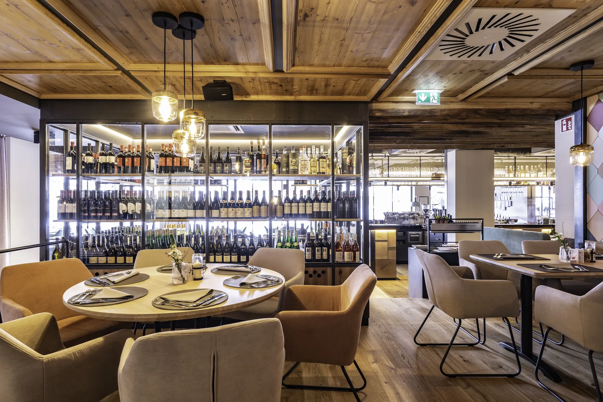 Gastraum des Restaurant Freestyle in Kaprun mit gedecktem Rundtisch, beigen Samtsesseln und einem großen beleuchteten Weinregal unter einer rustikalen Holzbalkendecke.