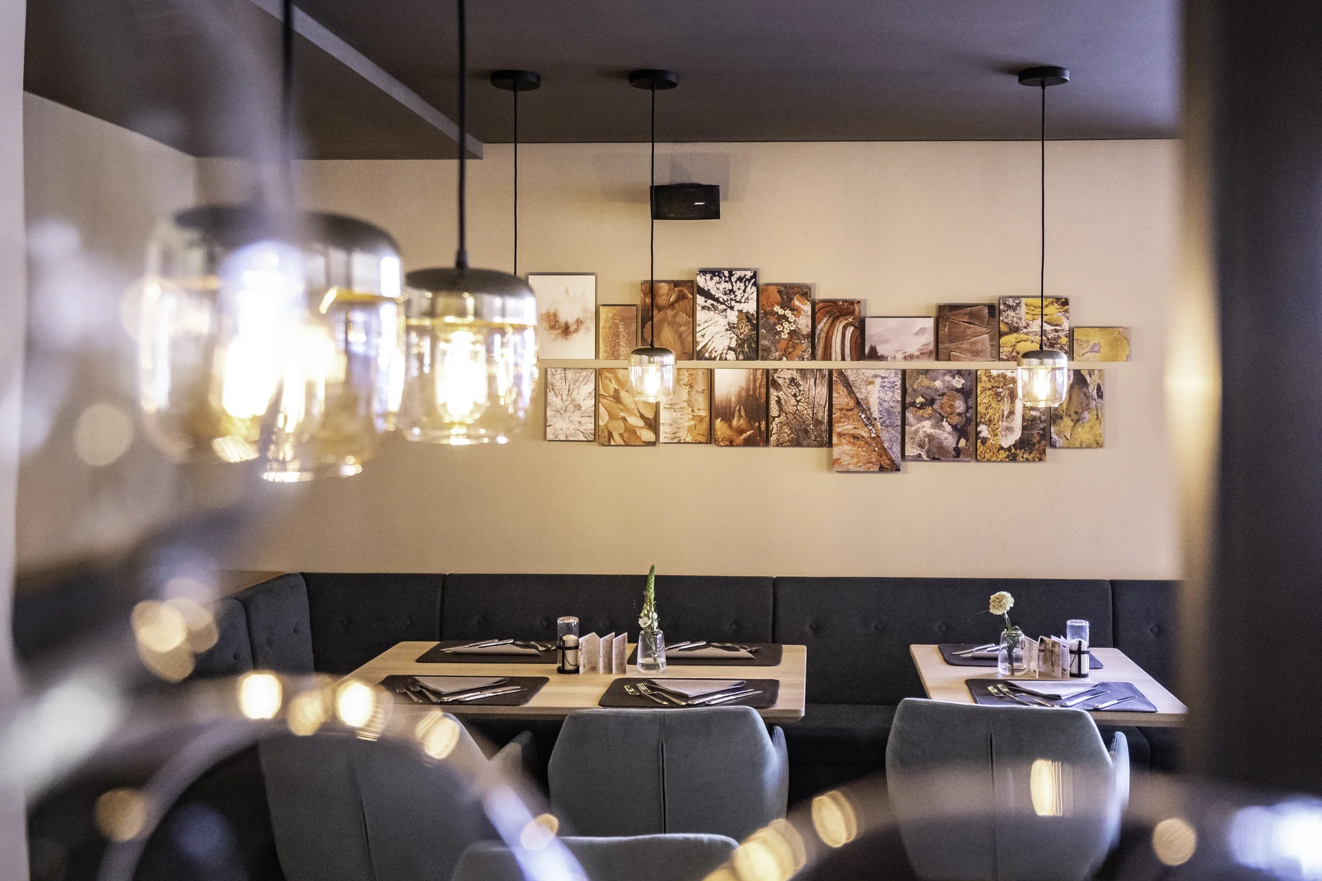 Sitzbereich im Restaurant Freestyle in Kaprun mit dunkler Samtbank, grauen Sesseln und gedeckten Tischen, dahinter eine lange Fotocollage aus Naturmotiven an der Wand, gerahmt von goldenen Glaspendelleuchten.