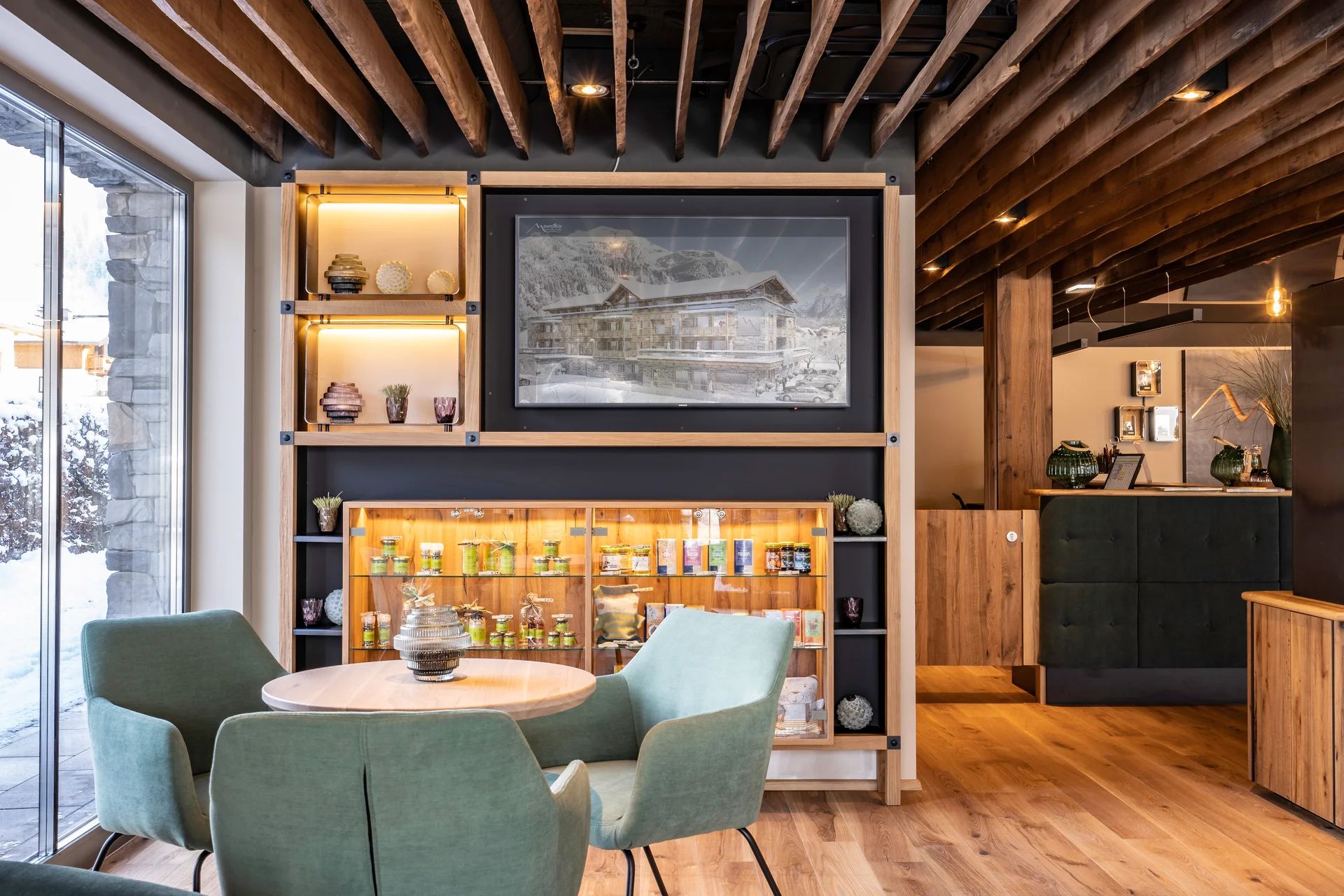 Lobby des Mountain Hotel Luis in Kaprun mit mintgrünen Samtsesseln, beleuchtetem Holzregal mit Produkten, einem winterlichen Hotelgemälde und dem Empfangsbereich im Hintergrund.