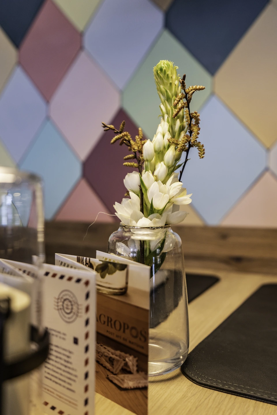 Tischdetail im Restaurant Freestyle in Kaprun mit einer Glasvase mit weißen Blüten und Weidenkätzchen vor der markanten bunten Rautenfliesenwand.