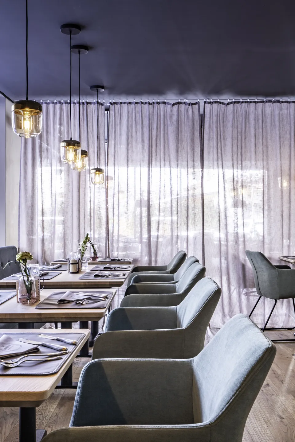 Sitzreihe im Restaurant Freestyle in Kaprun mit mintgrünen Samtsesseln an gedeckten Holztischen, goldenen Glaspendelleuchten und lichten Leinenvorhängen vor den Panoramafenstern.