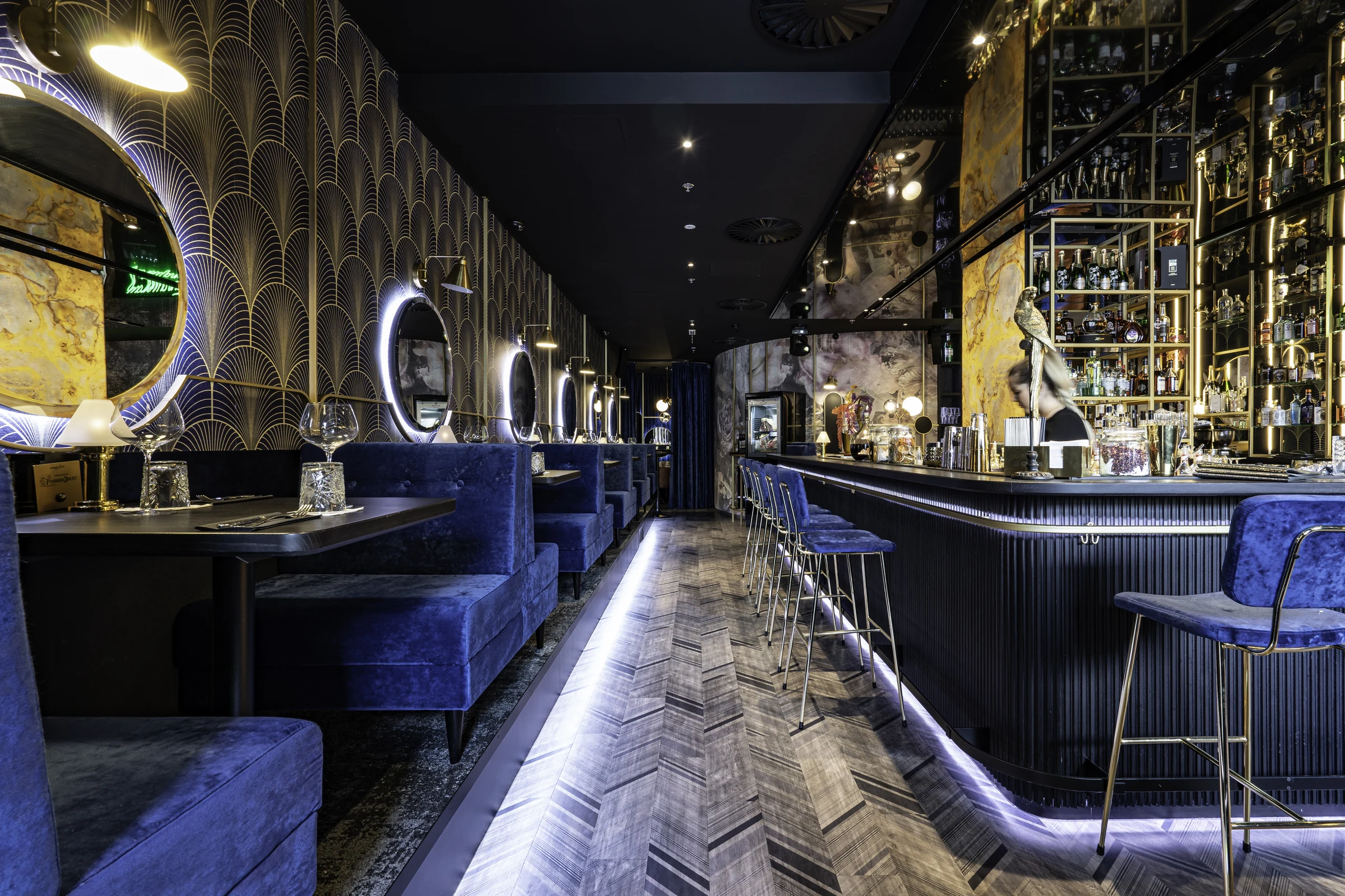 Gesamtansicht der Nuba Bar in Linz mit langer blauer Samtbank, Art-Déco-Tapete mit Goldmuster, beleuchteten Rundspiegel und einem imposanten Spirituosenregal entlang des Fischgrätbodens.