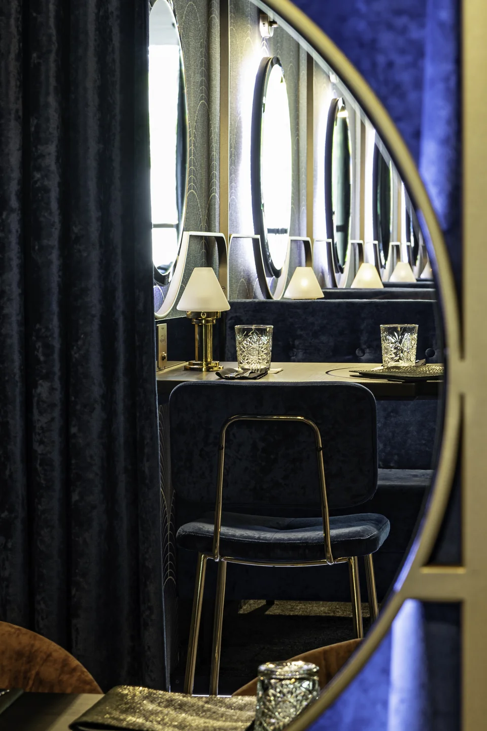 Blick durch einen goldgerahmten Rundspiegel auf die endlos wirkende Spiegelreihe der Nuba Bar in Linz mit blauer Samtbank, goldenen Tischlämpchen und Kristallgläsern.