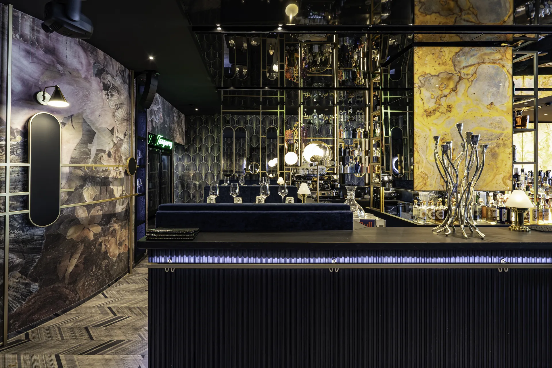 Blick hinter den Bartresen der Nuba Bar in Linz mit goldenem Spirituosenregal, gelbem Marmorpaneel, Art-Déco-Tapete und silberner Kerzenleuchterskulptur auf dem schwarzen Tresen.
